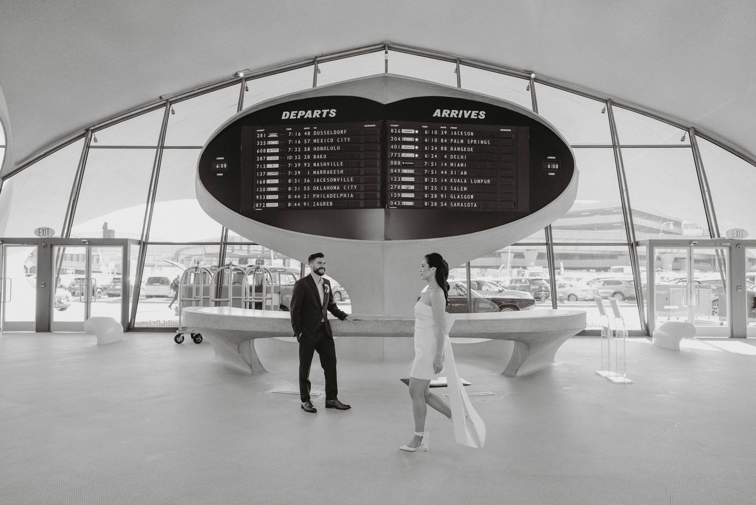 Bride and groom stand apart in black and white photo beneath departures and arrivals board.