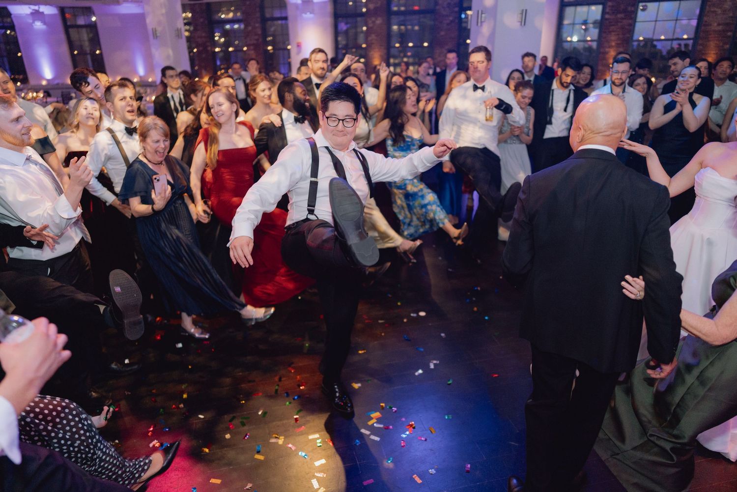Energetic wedding guest dancing at a lively reception, surrounded by celebrating guests on a confetti-covered dance floor.