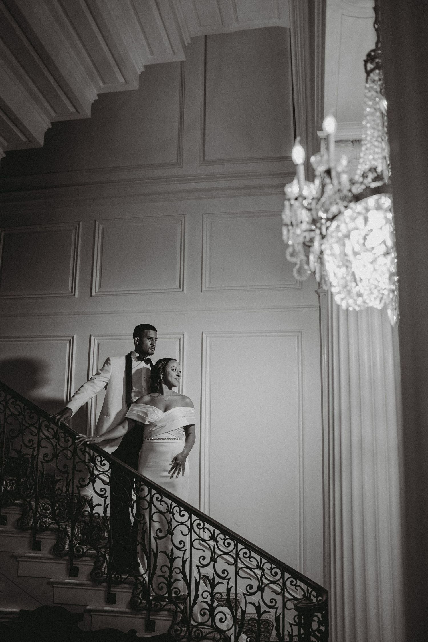 Elegant black and white photo of bride and groom on ornate staircase beneath crystal chandelier.