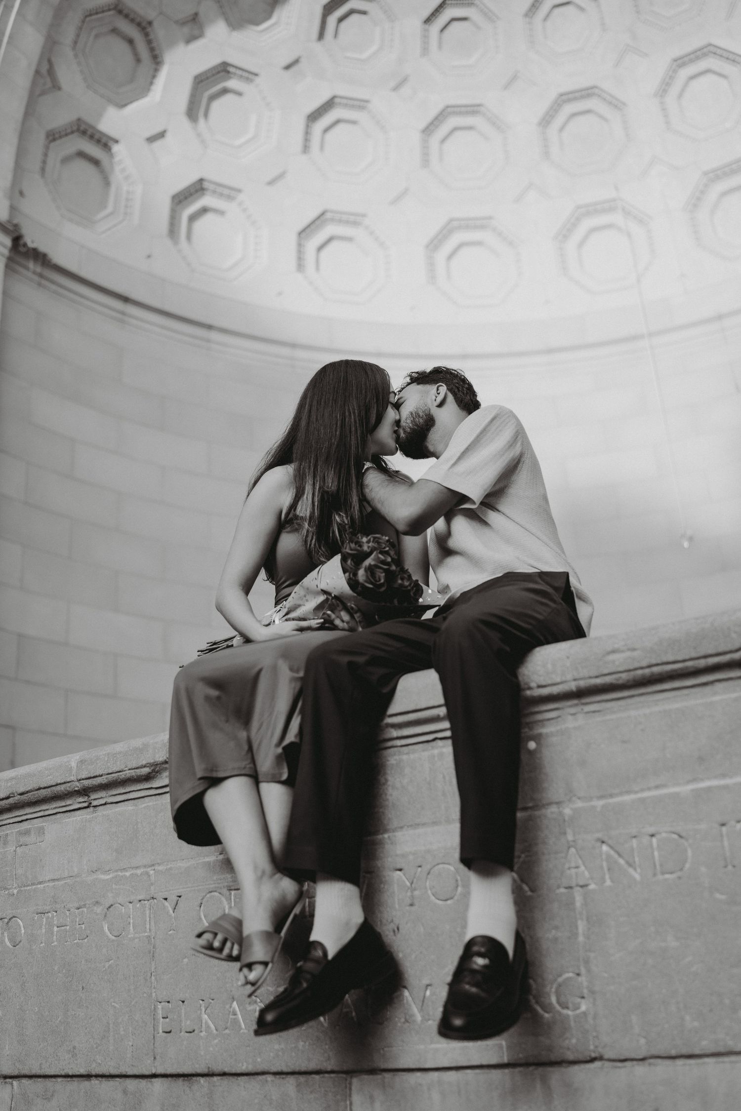 An engaged couple shares a kiss while seated on a stone ledge beneath an ornate domed ceiling in black and white.