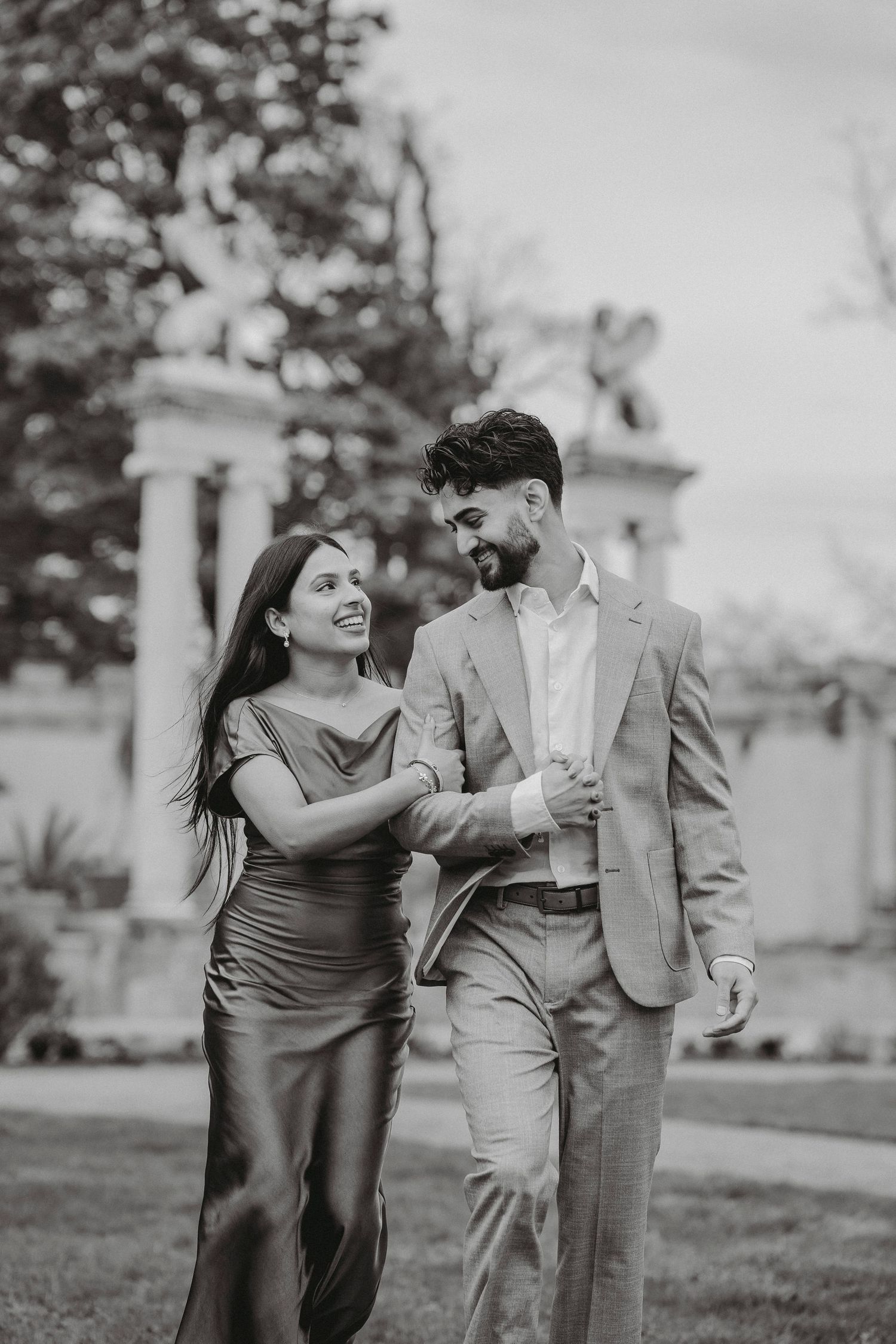 Black and white photo of laughing couple in formal wear strolling through a garden together.