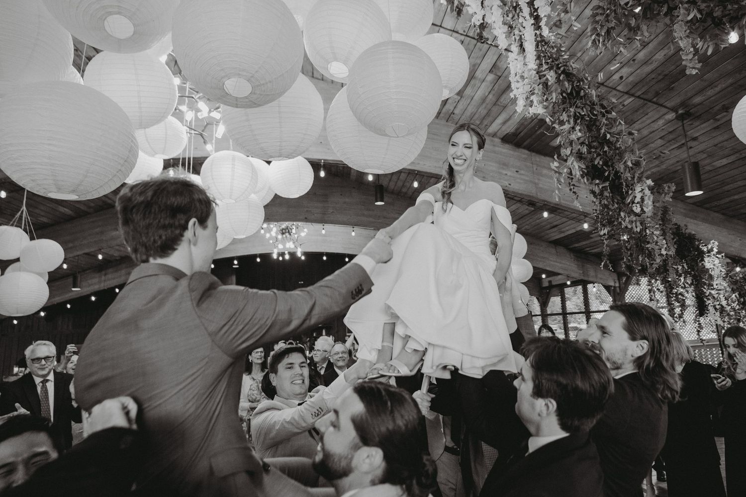 Black and white photo of bride lifted by guests during lively wedding reception dance celebration.