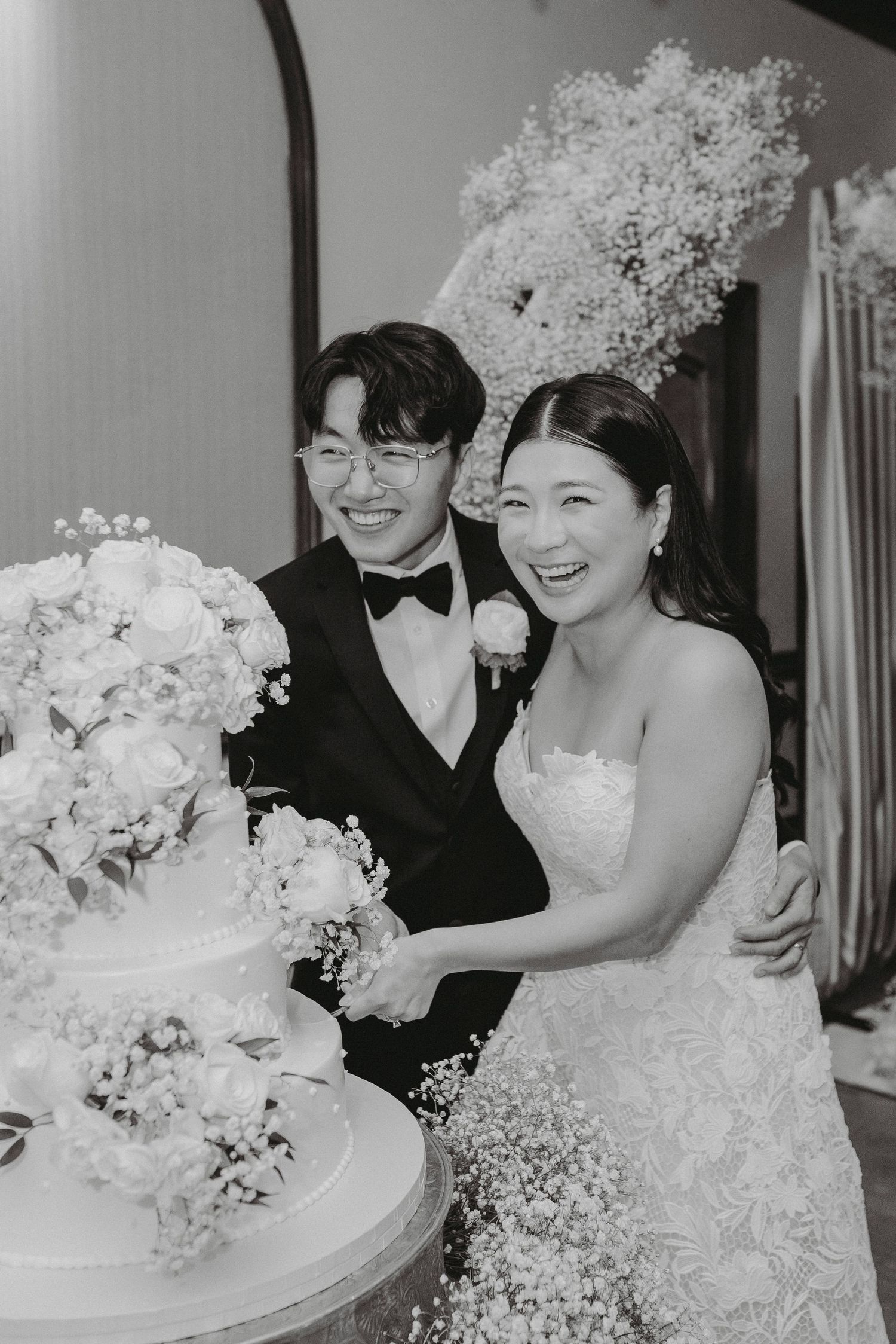 Laughing bride and groom cut elegant floral wedding cake surrounded by baby's breath in black and white photo.