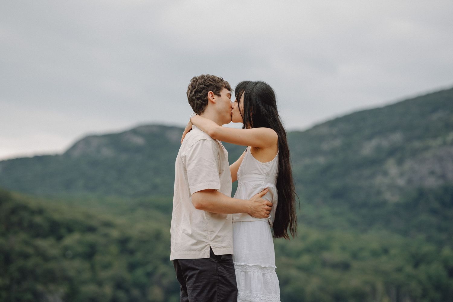 Couple in casual summer clothes embrace and kiss in front of forested green mountains under an overcast sky.