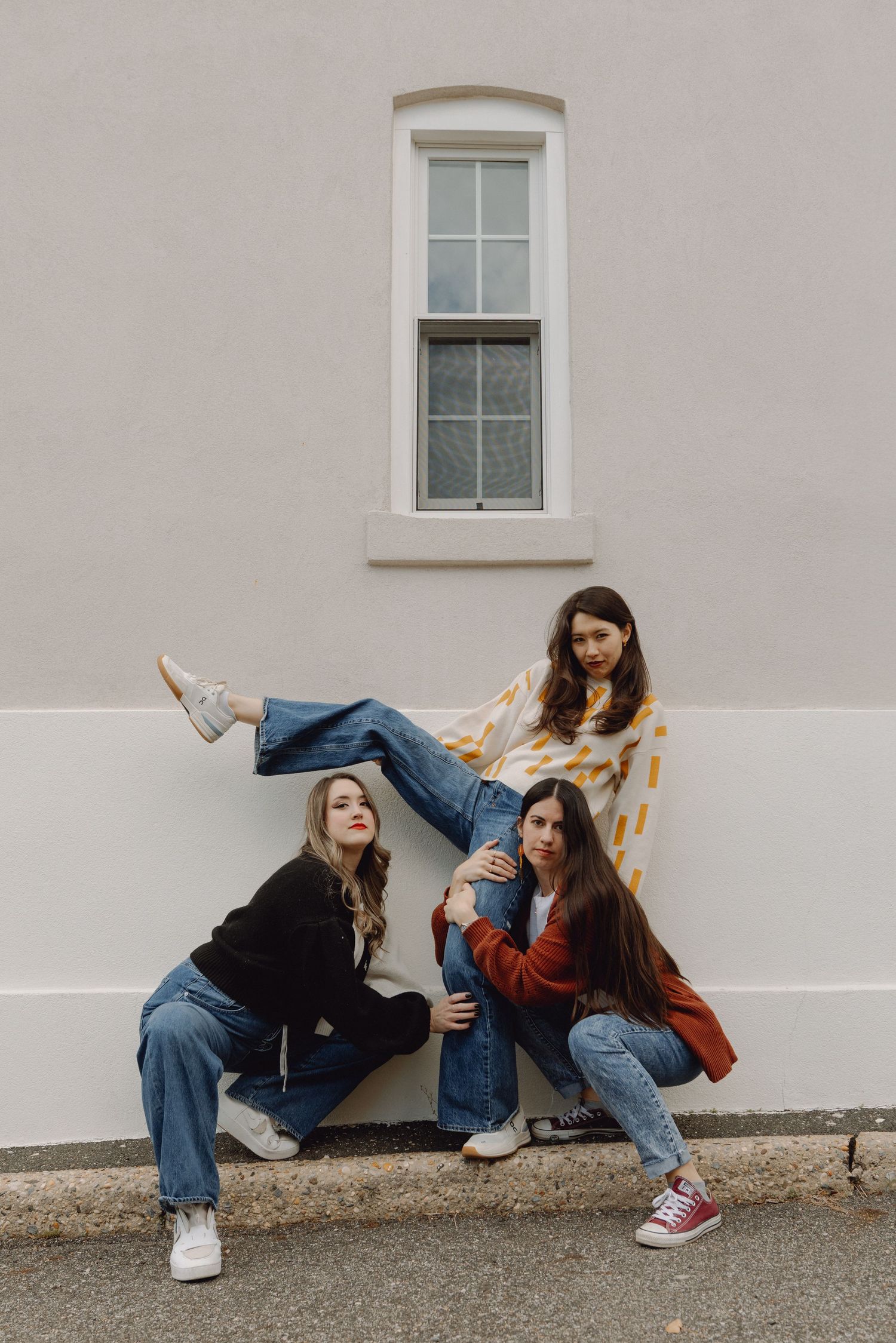 Three young women pose playfully against a building wall wearing jeans and colorful tops.