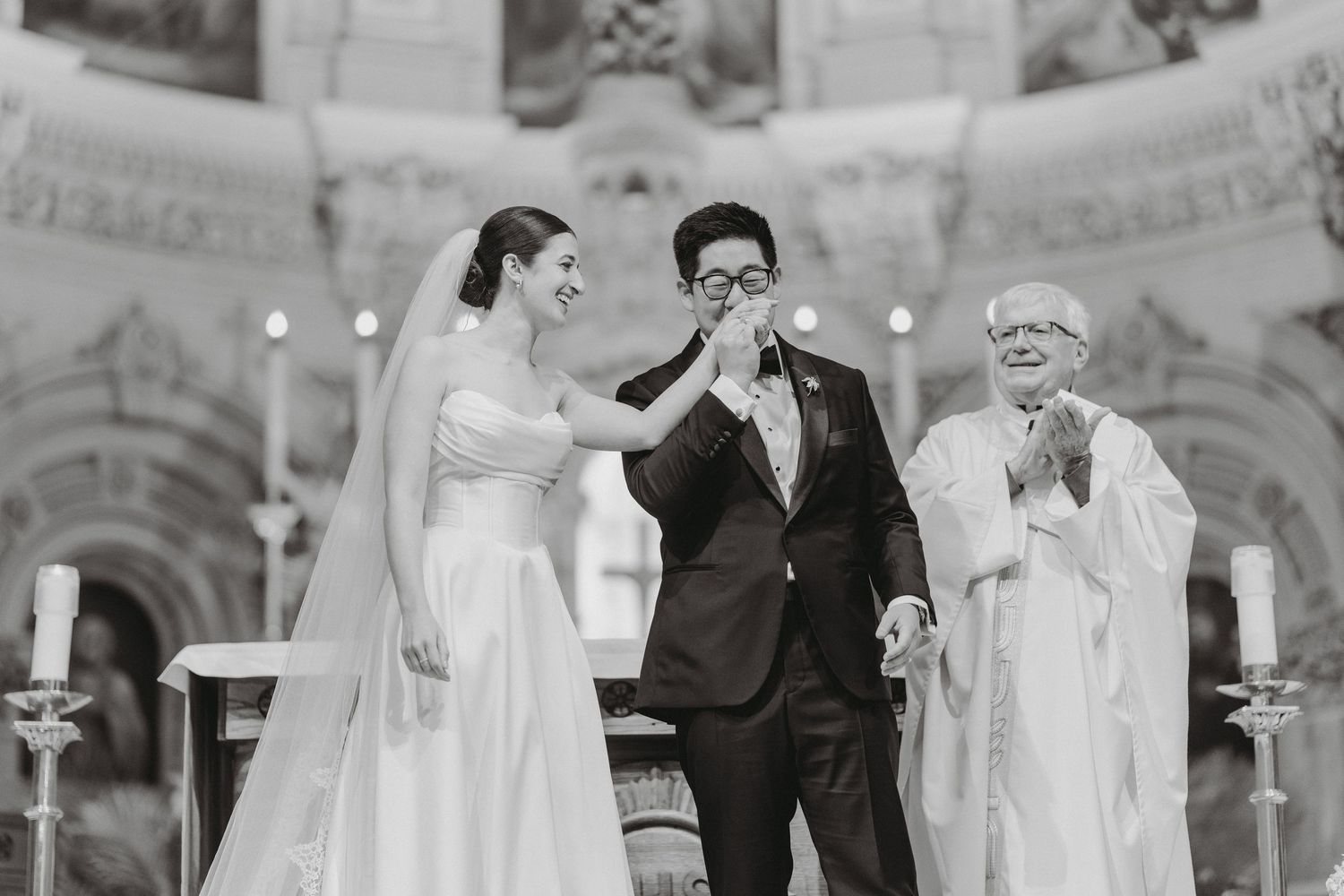 Groom kissing bride's hand at altar while priest applauds in black and white wedding photo.