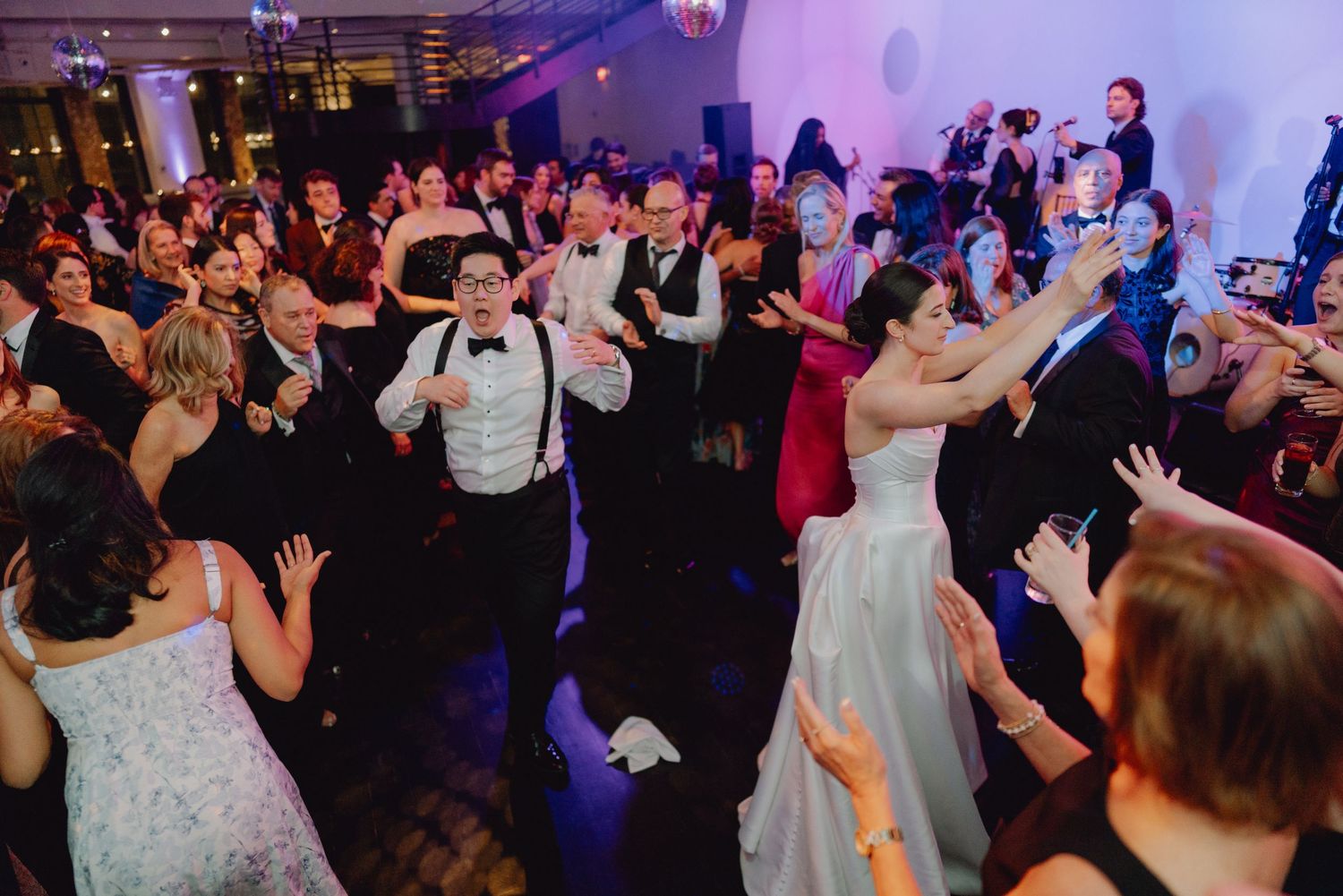Bride and groom celebrate with guests on a crowded, energetic wedding reception dance floor.