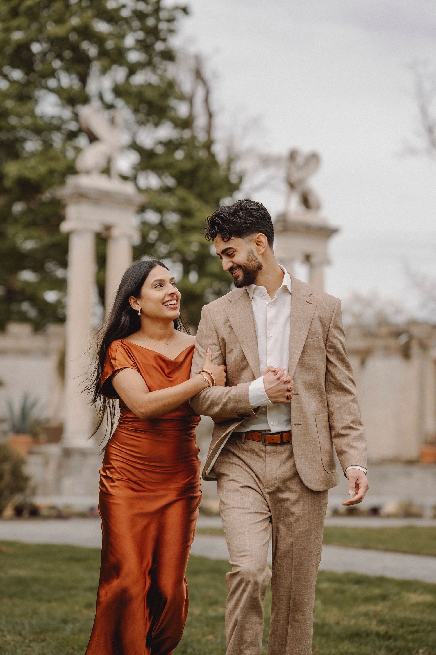 Laughing couple walking arm in arm past decorative columns with angel sculptures in a garden.