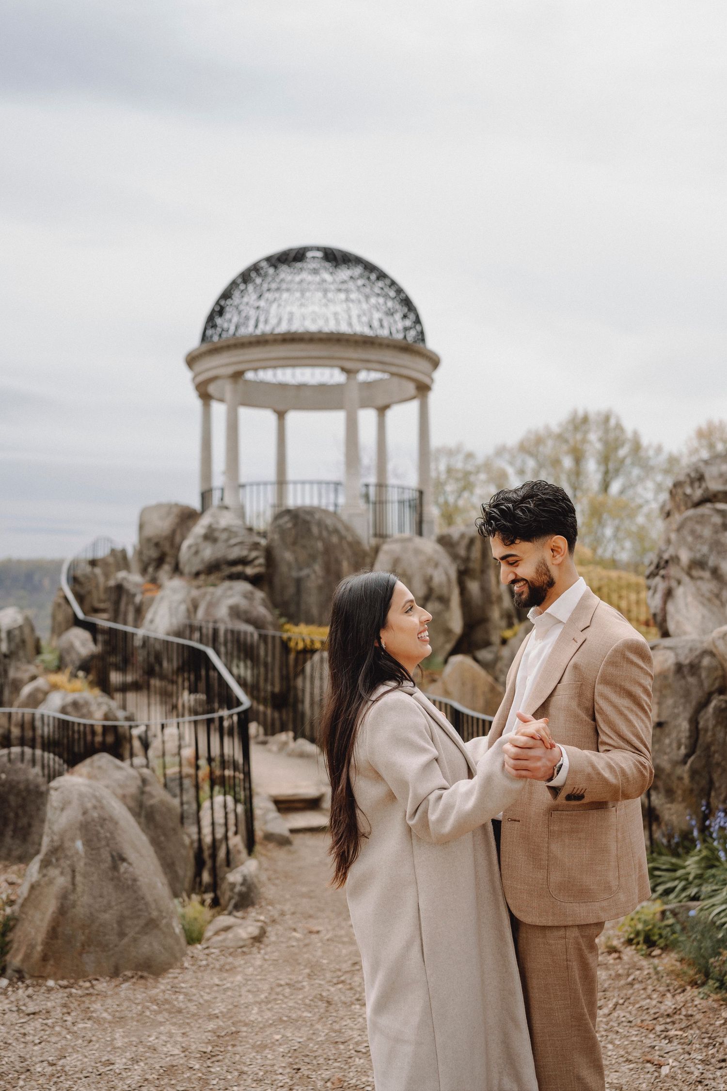 Couple holding hands facing each other with ornate glass dome gazebo on rocky hilltop behind them.