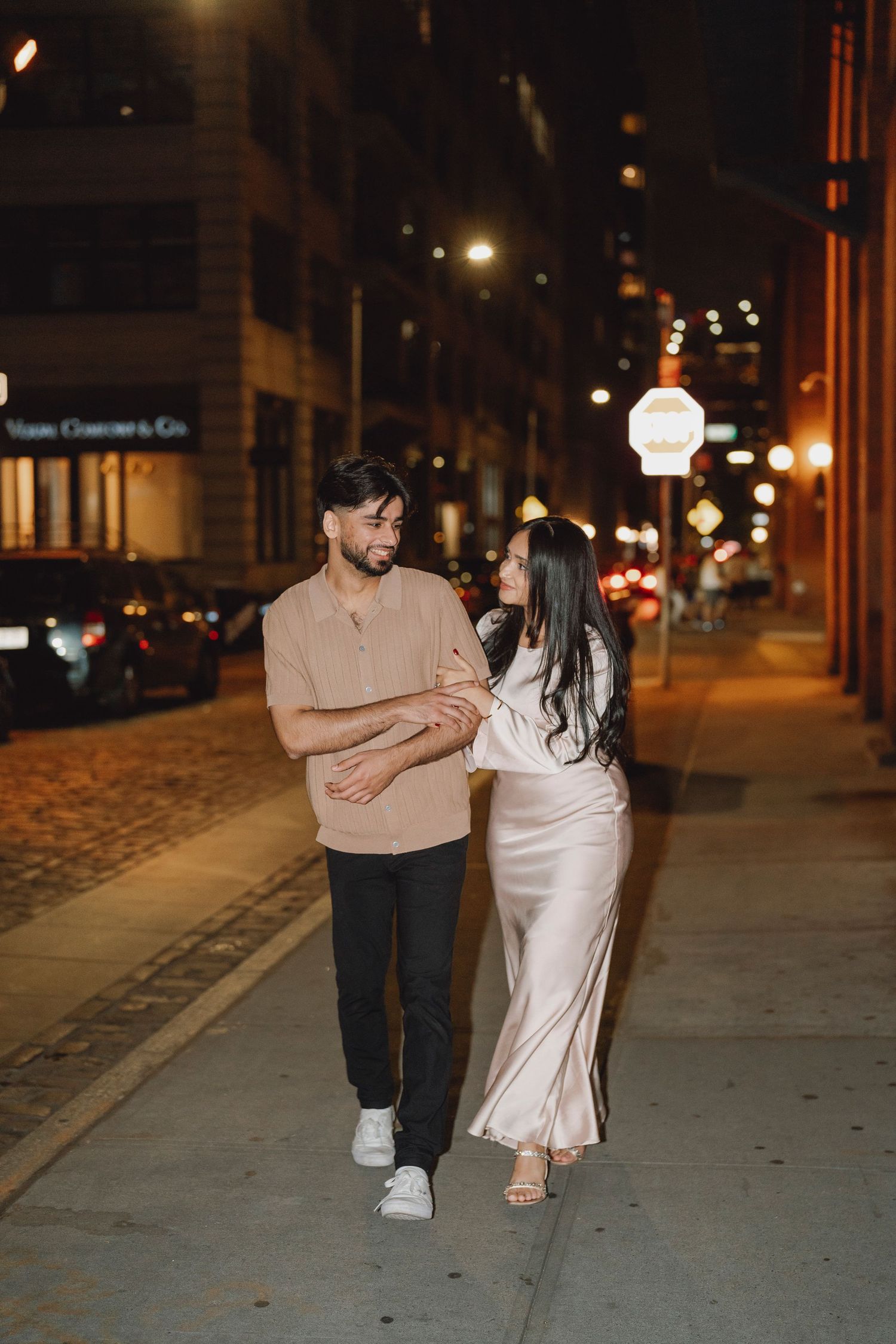 Smiling couple walking arm in arm along a city sidewalk at night, he in a tan shirt and she in a silk champagne dress.