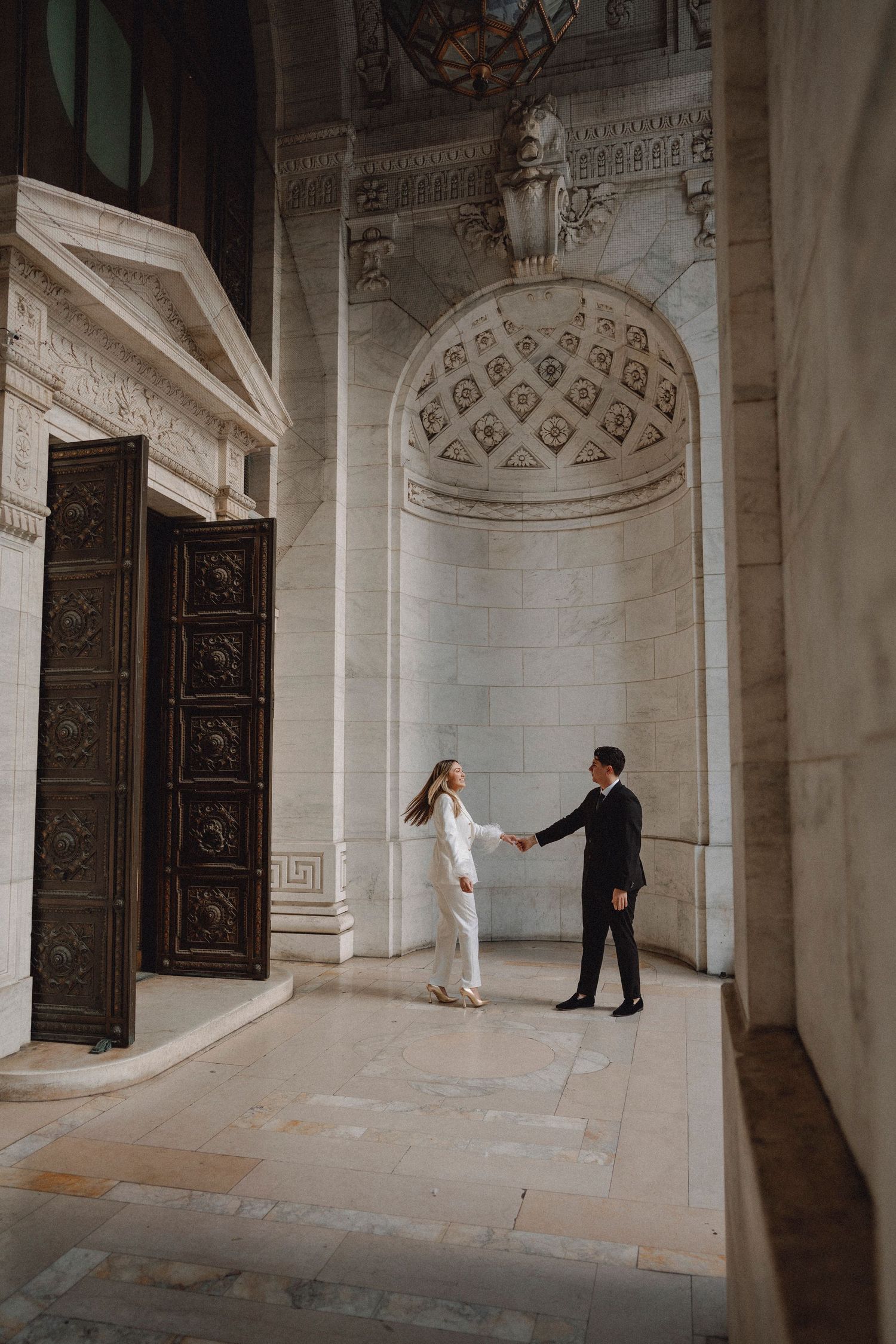 Couple holding hands in an ornate marble building with decorative bronze doors.