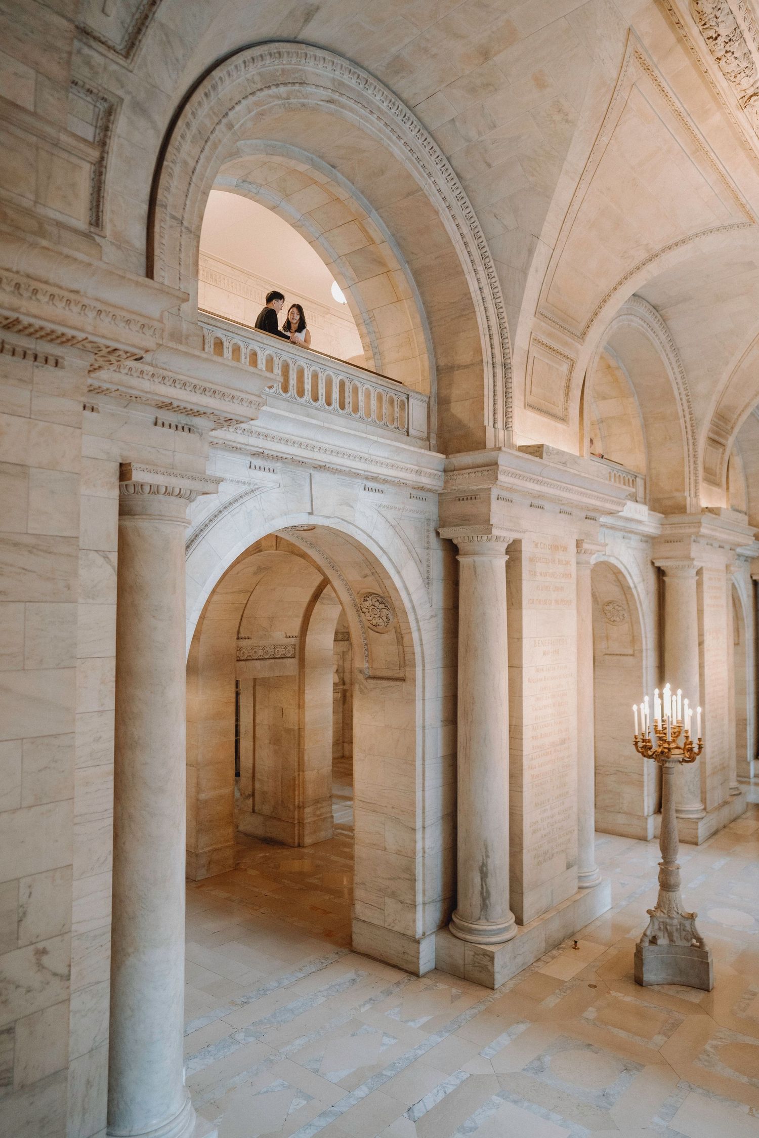 Couple standing on an upper balcony overlooking a grand marble hall with vaulted arches and a golden candelabra.