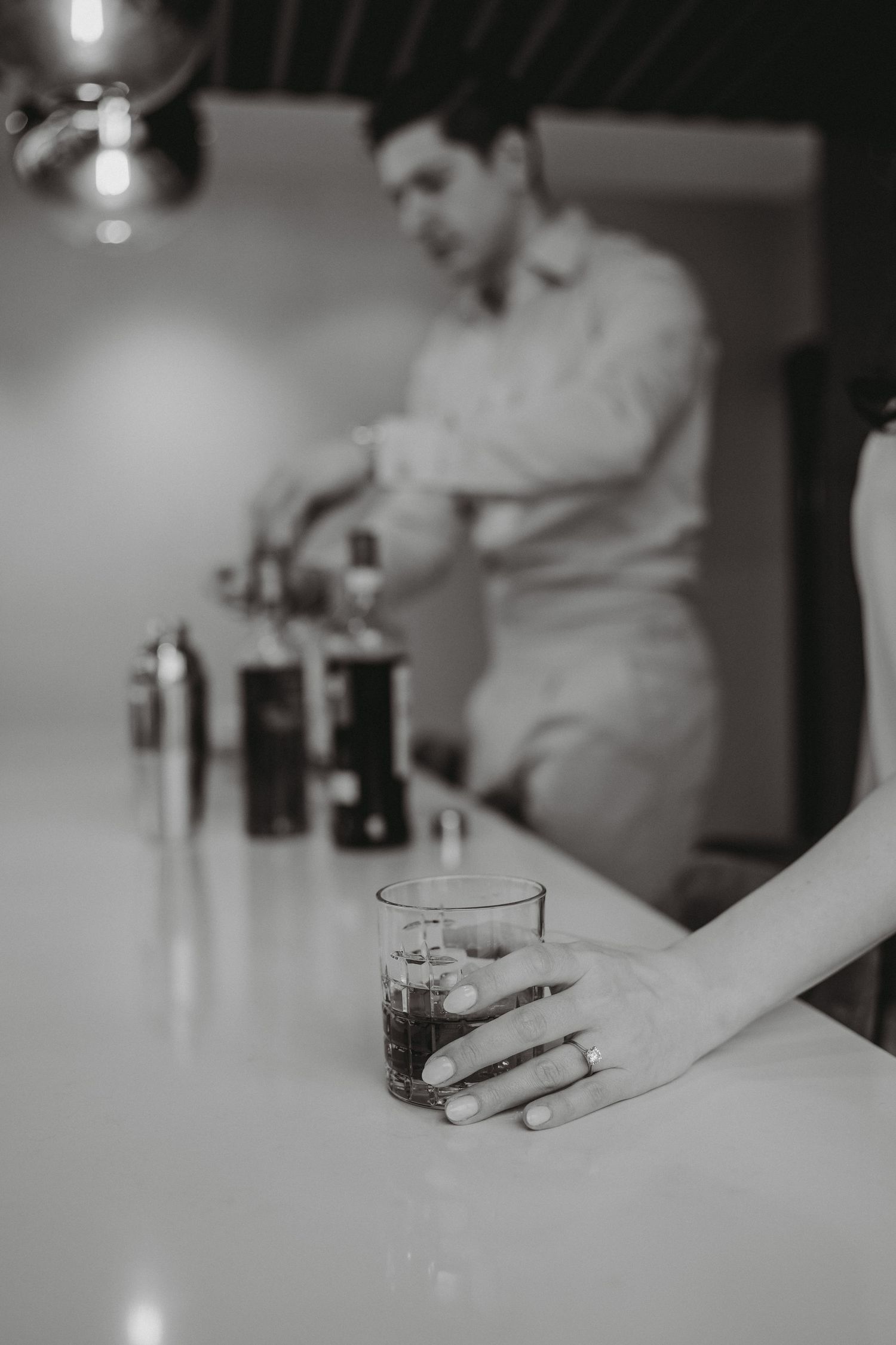 Black and white close-up of a hand with engagement ring holding a drink at a bar.