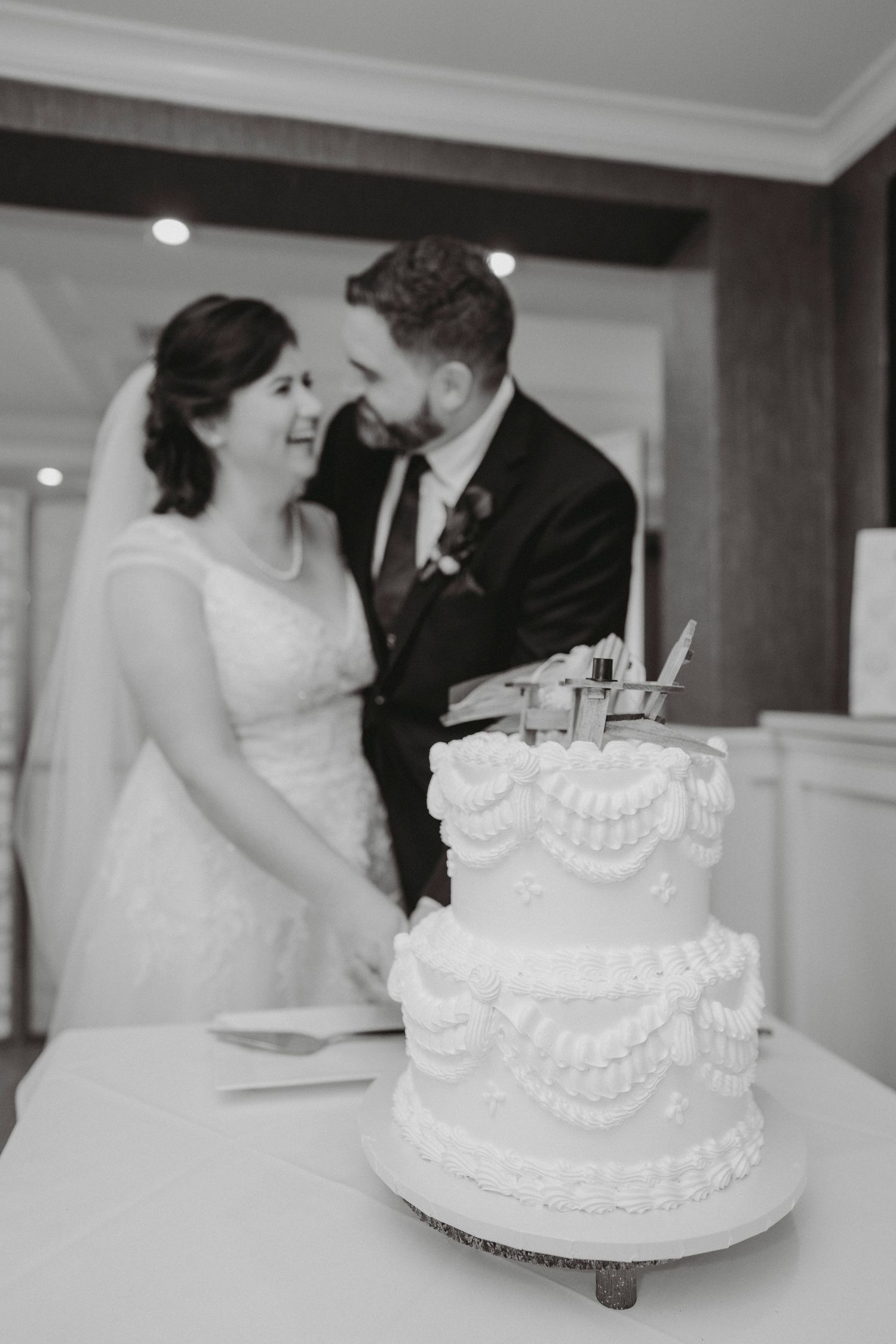 Bride and groom share intimate moment cutting elegant two-tier wedding cake in black and white photo.