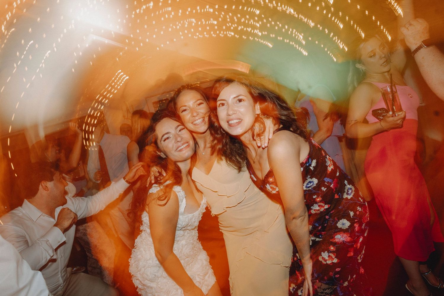Three smiling women pose together at a wedding reception surrounded by twinkling lights and guests.