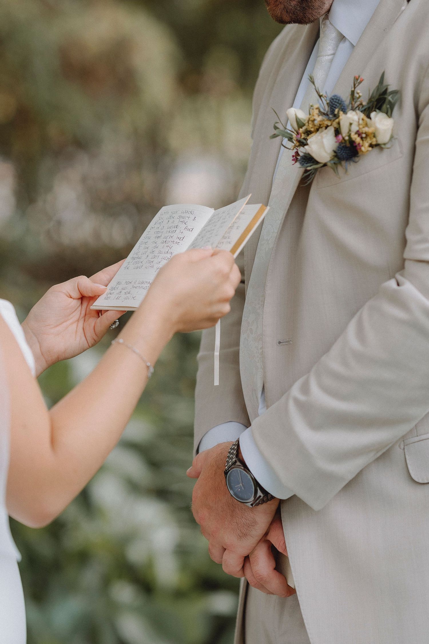 Groom in a beige suit with boutonniere listening as bride reads wedding vows from a small notebook.