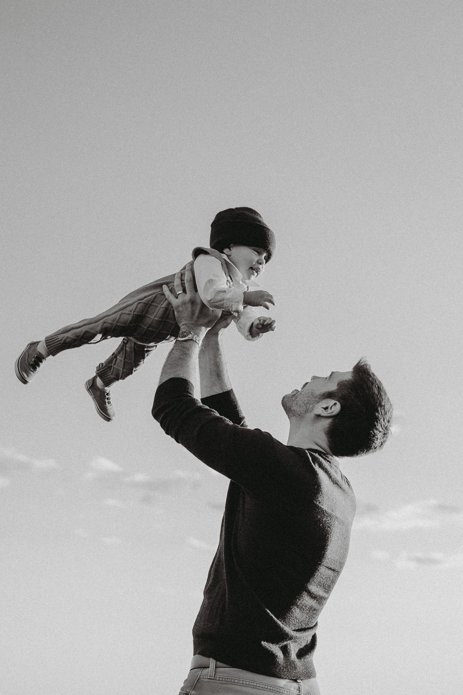 Black and white photo of a father joyfully lifting his baby high in the air against a cloudy sky.