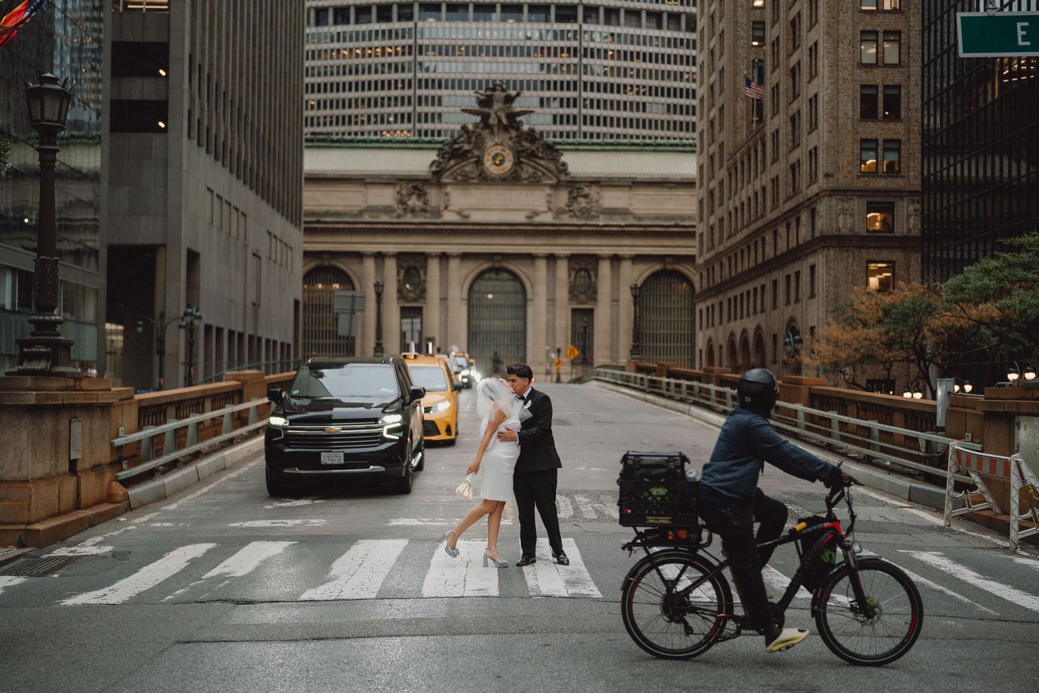 Bride and groom crossing street at crosswalk near Grand Central Terminal in New York City with cyclist passing by.