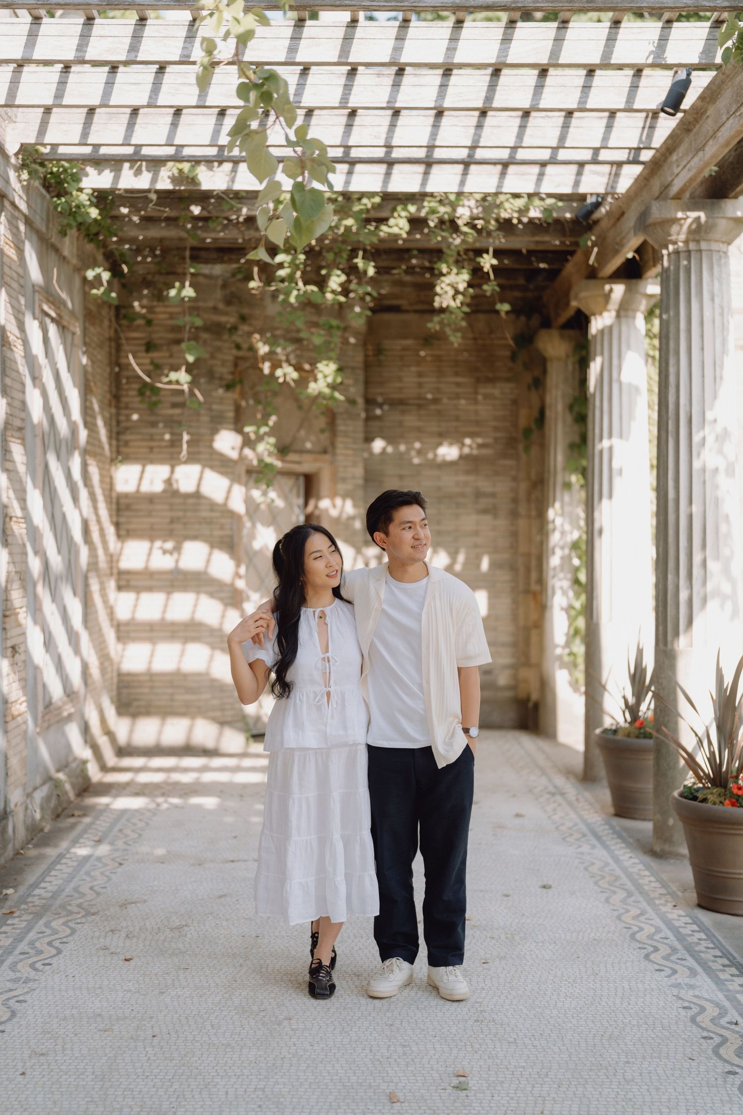 Smiling couple in white outfits embracing under a vine-covered pergola with sunlight streaming across a stone columned walkway.