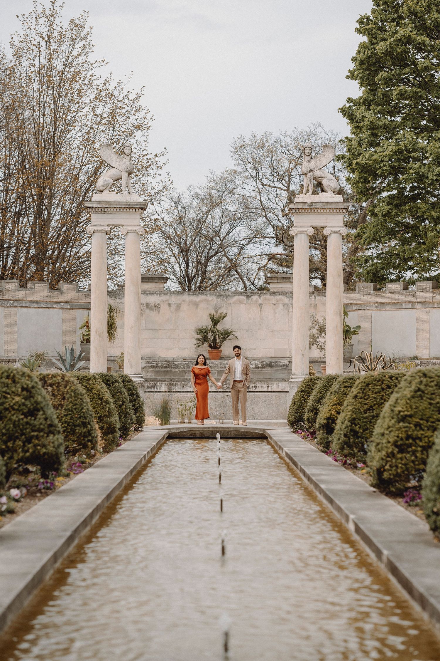 Couple holding hands beside a long reflecting pool framed by columns with griffin statues.