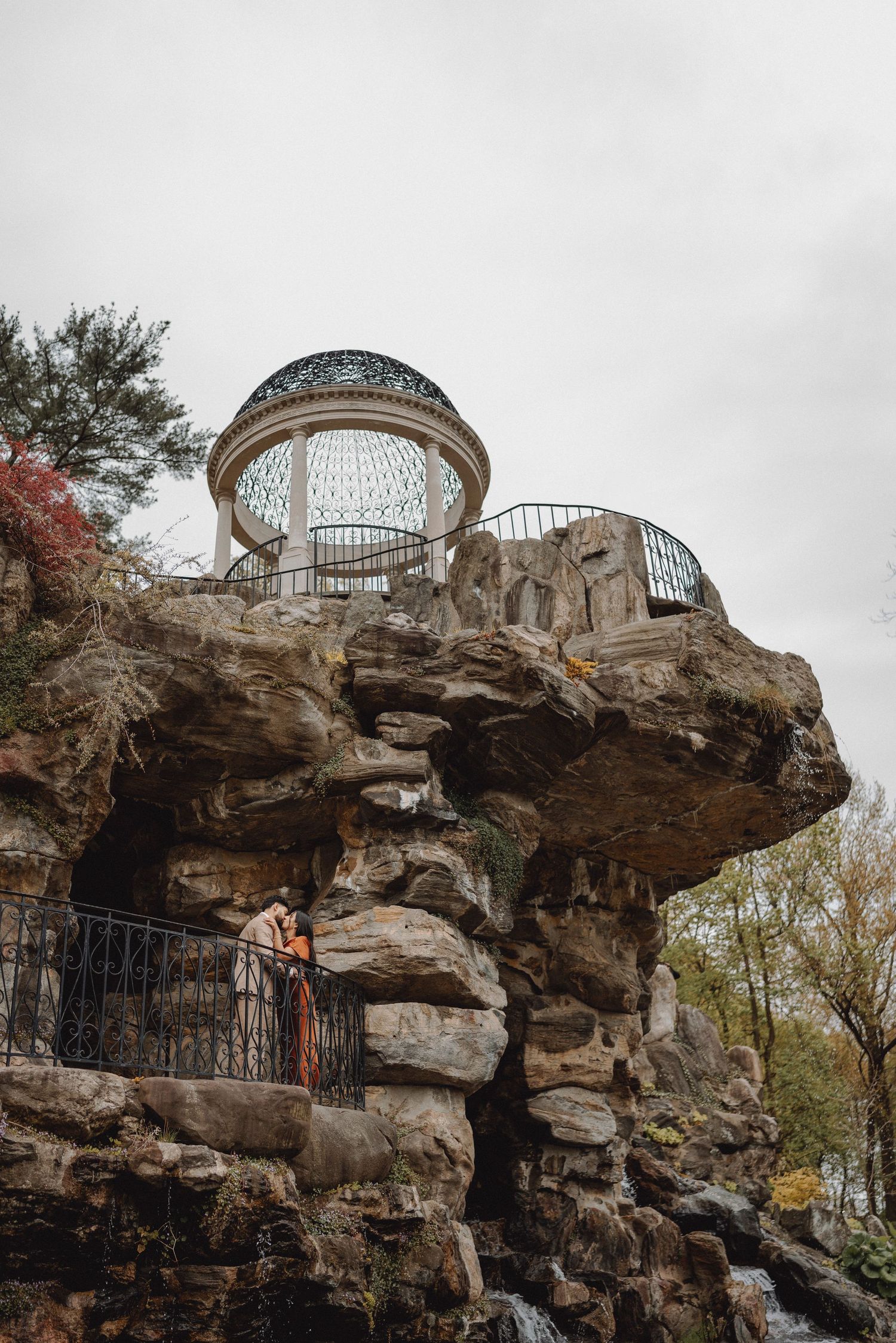 Dramatic rocky landscape with ornate glass dome gazebo perched on top, couple visible on lower rock ledge.