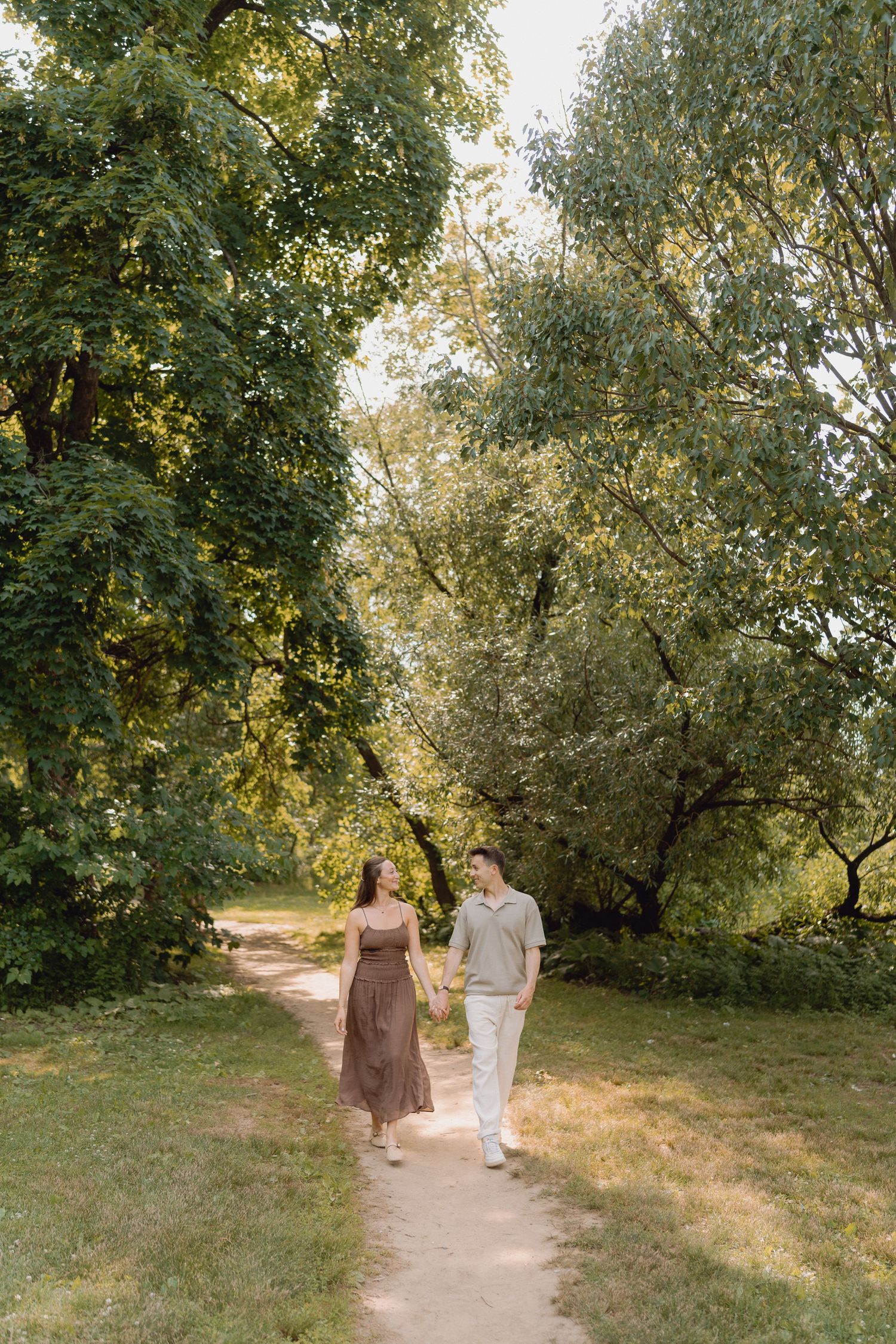 Couple holding hands and walking along a dirt path through a sunlit green park surrounded by lush trees.