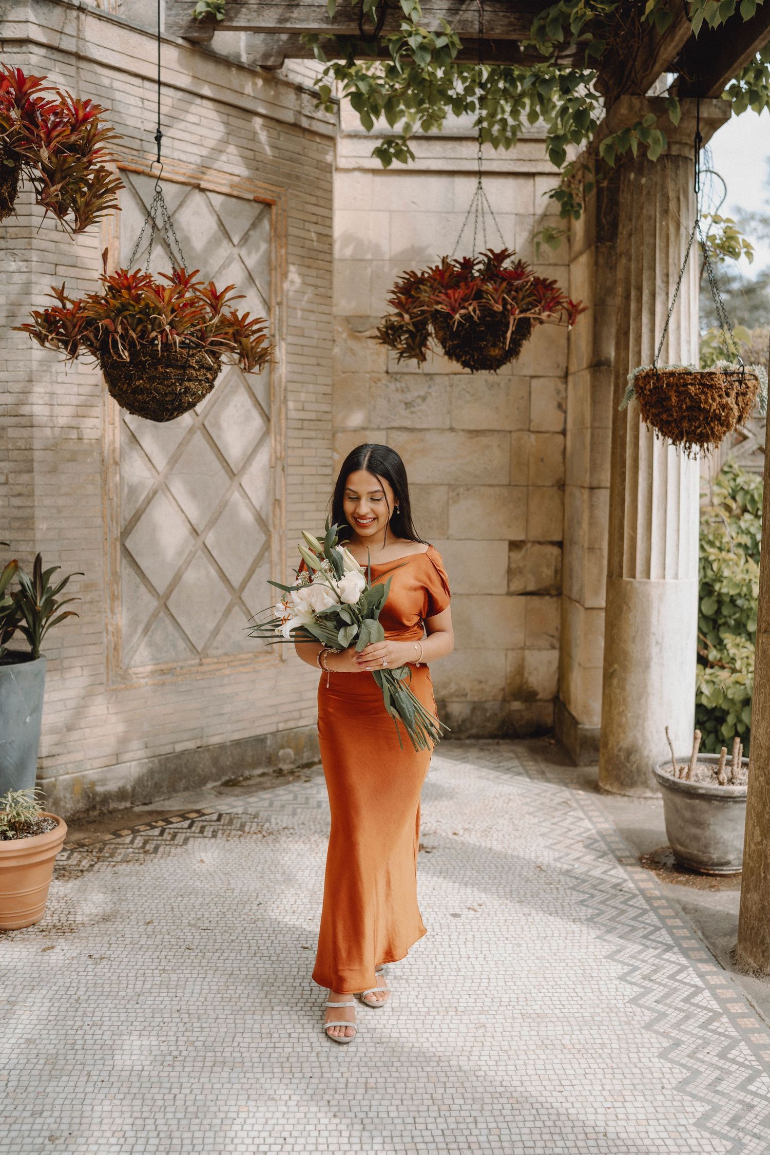 Woman in orange satin dress walks through a sunlit pergola holding a tropical floral bouquet.