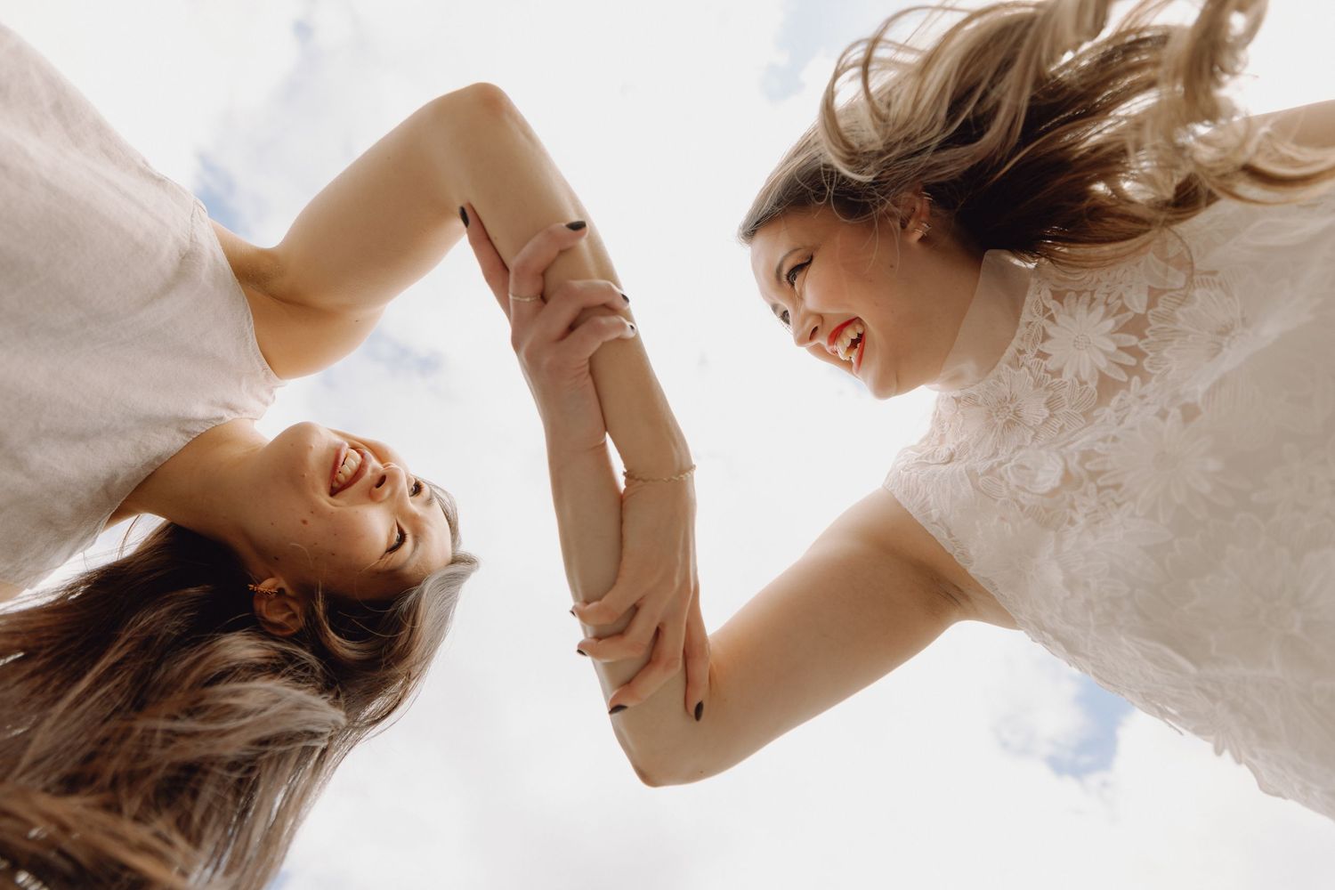 Two smiling women hold hands viewed from below against a bright blue sky.