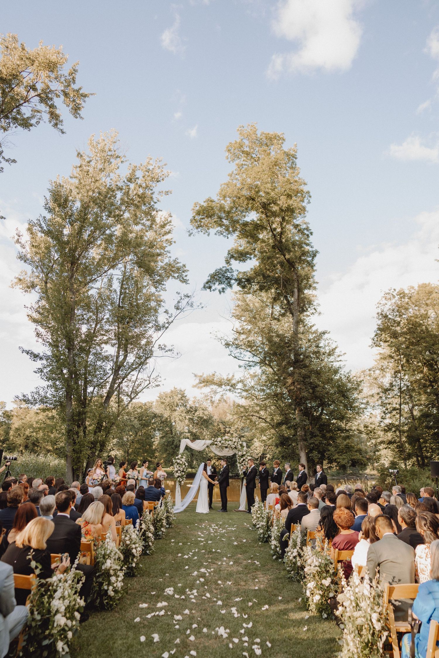 Elegant outdoor wedding ceremony with white floral aisle and arch surrounded by tall trees.