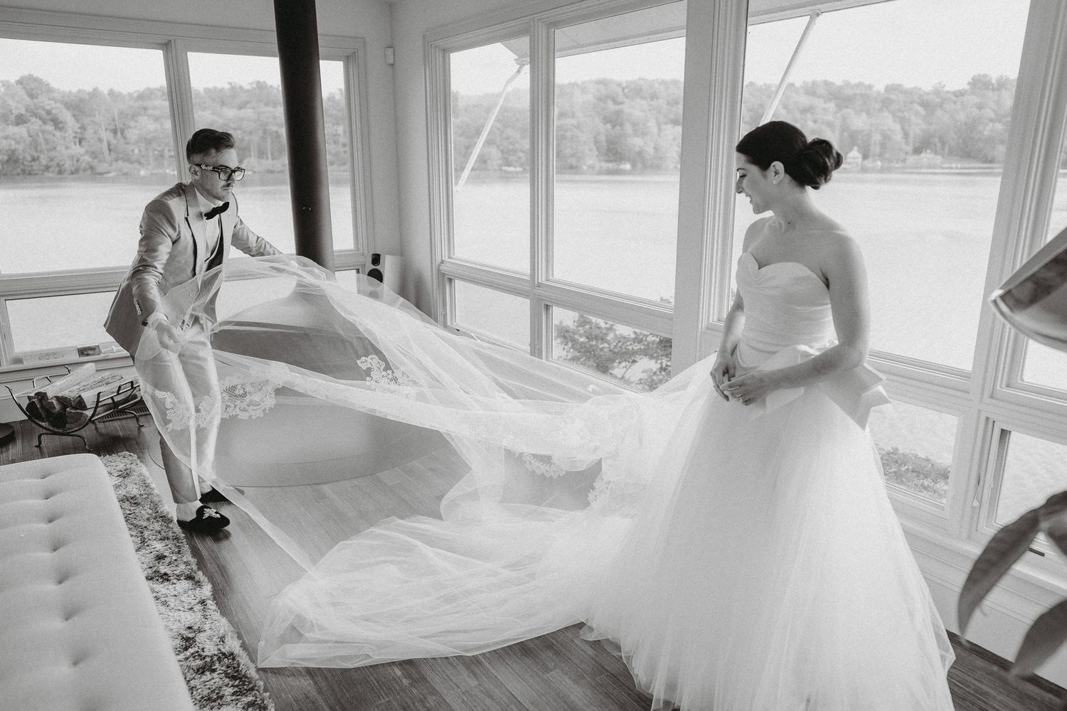 Black and white photo of groom spreading bride's cathedral veil in sunlit lakeside room.