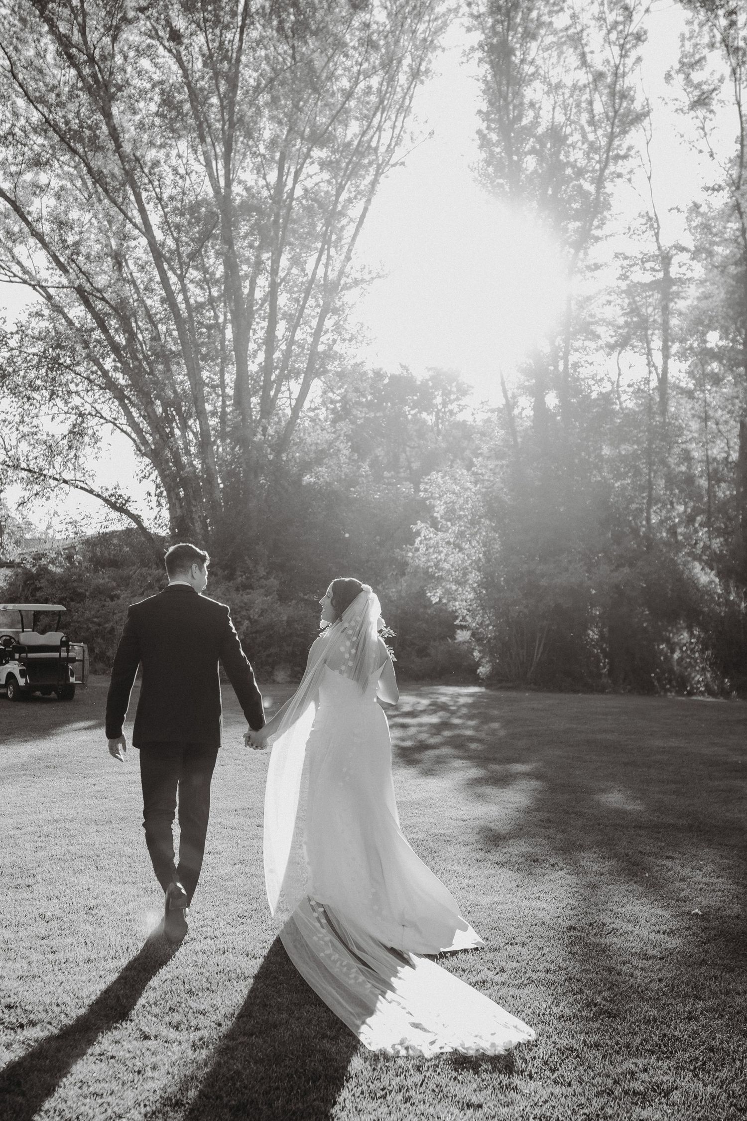 Black and white photo of bride and groom walking hand in hand through sunlit trees on their wedding day.