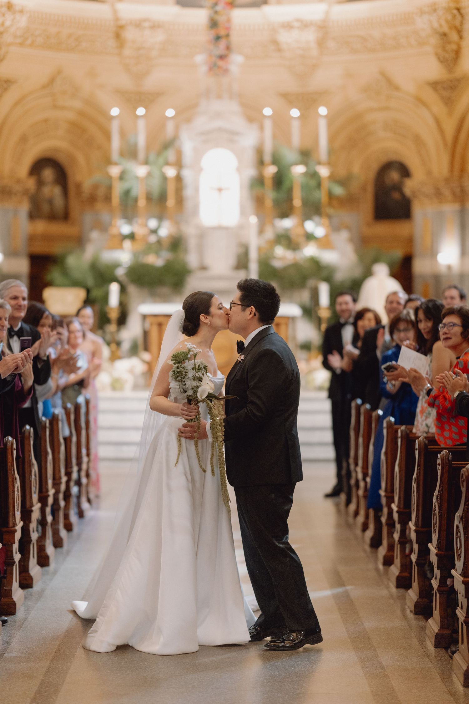 Bride and groom share first kiss walking down the aisle of an ornate golden Catholic church.