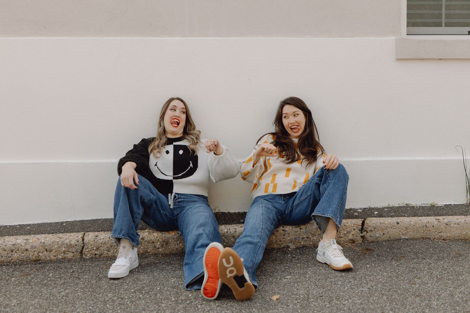 Two young women sitting against a white wall outdoors, wearing casual jeans and sneakers, laughing and posing playfully.
