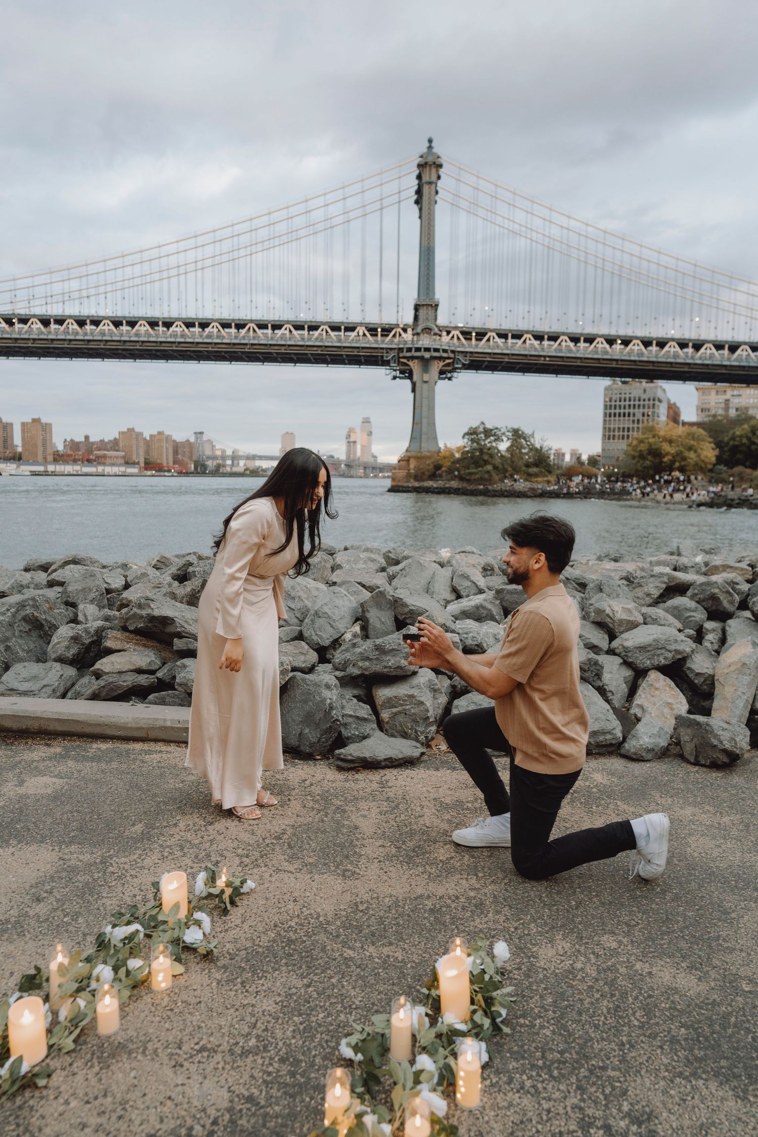 Man kneels with a ring during a candlelit proposal on a rocky waterfront as a woman watches, with a suspension bridge and skyline behind.