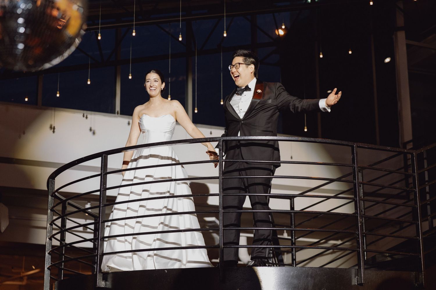 Bride and groom stand on spiral staircase balcony in elegant industrial wedding venue at night.