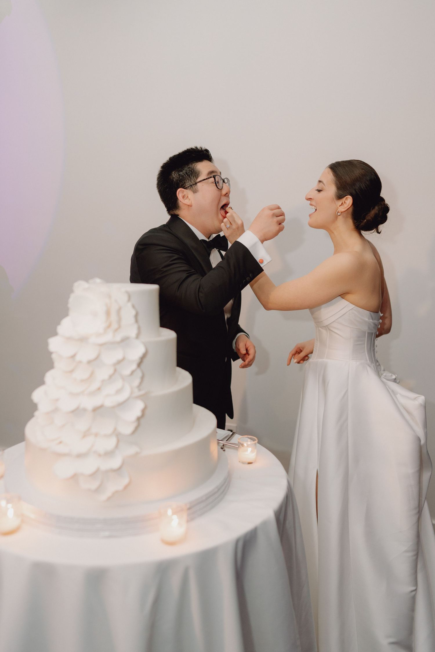 Smiling bride and groom feed each other wedding cake at their elegant white floral tiered cake table.