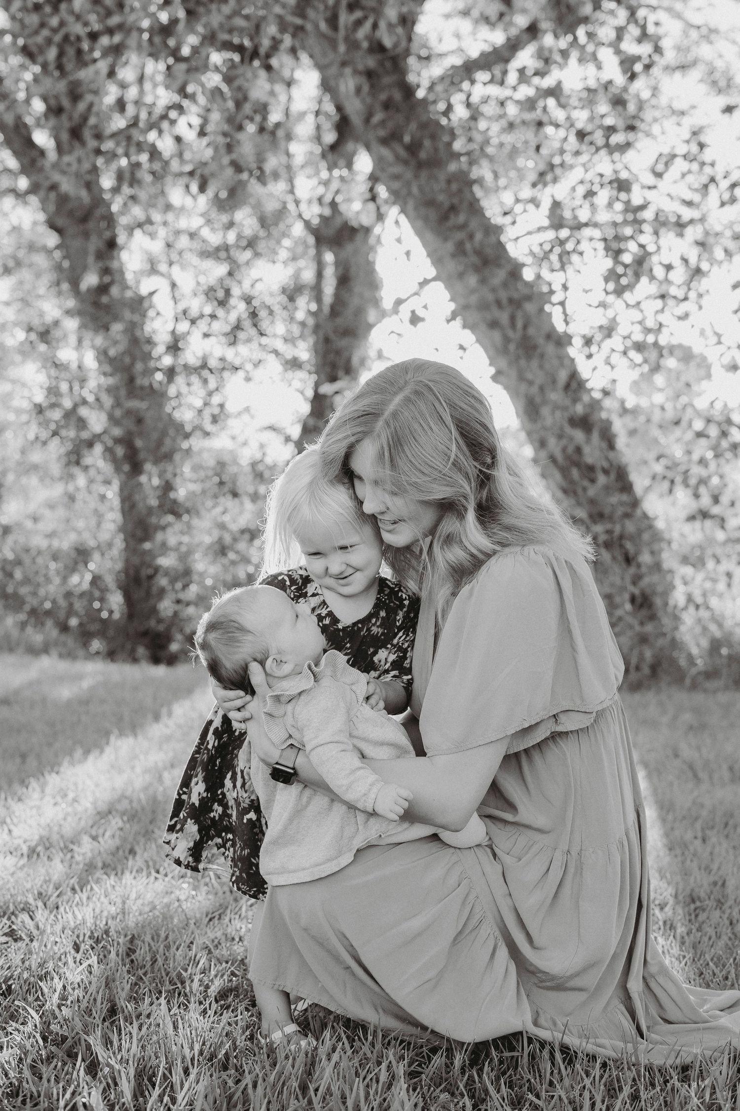 Mother kneeling outdoors with two young children under a tree in black and white.
