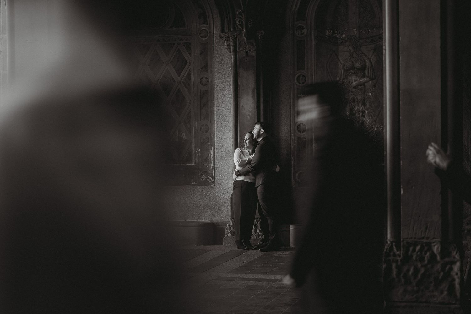 Black and white photo of a couple embracing in a dimly lit ornate interior hallway.