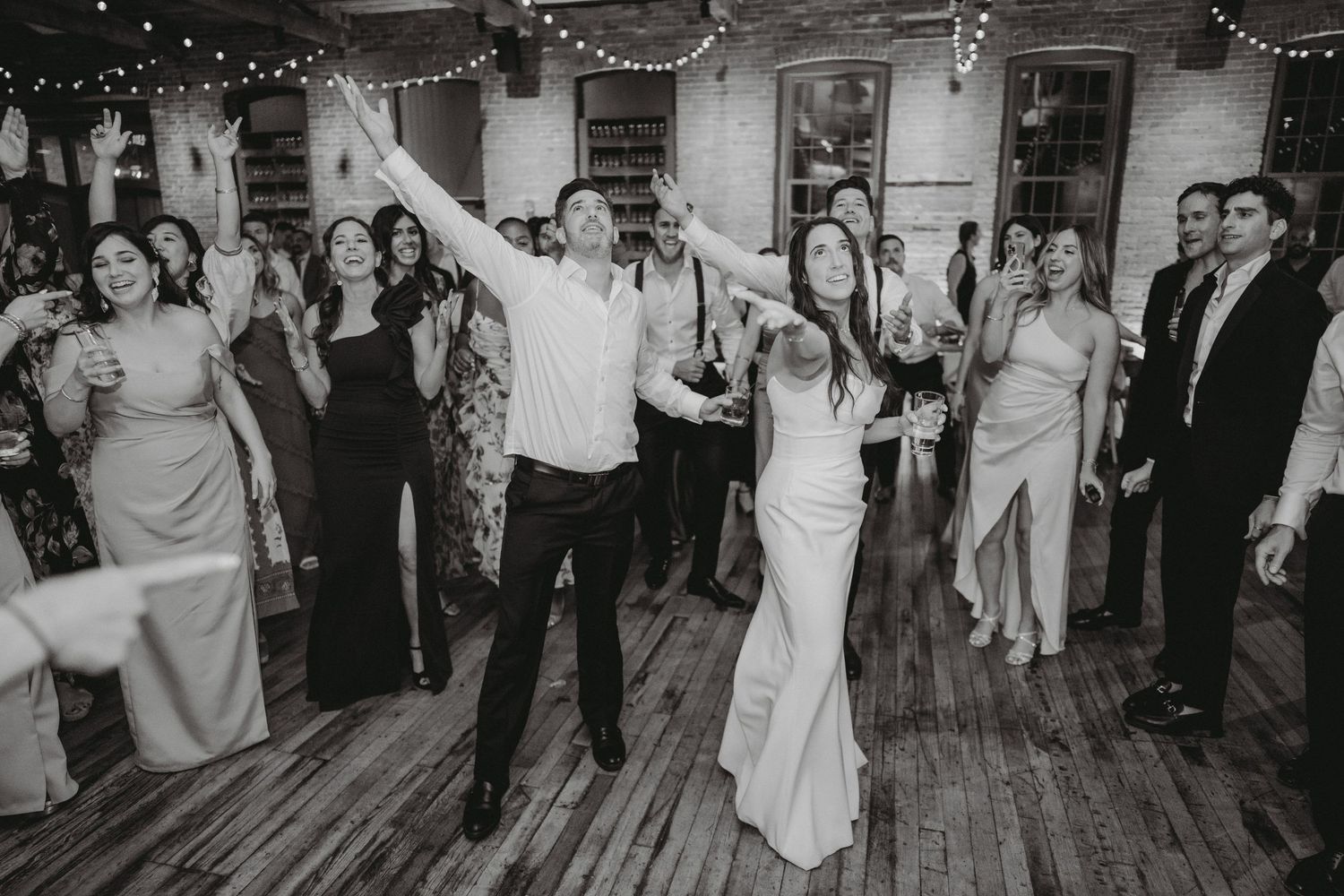 Bride and groom celebrate with raised arms surrounded by cheering guests at wedding reception.