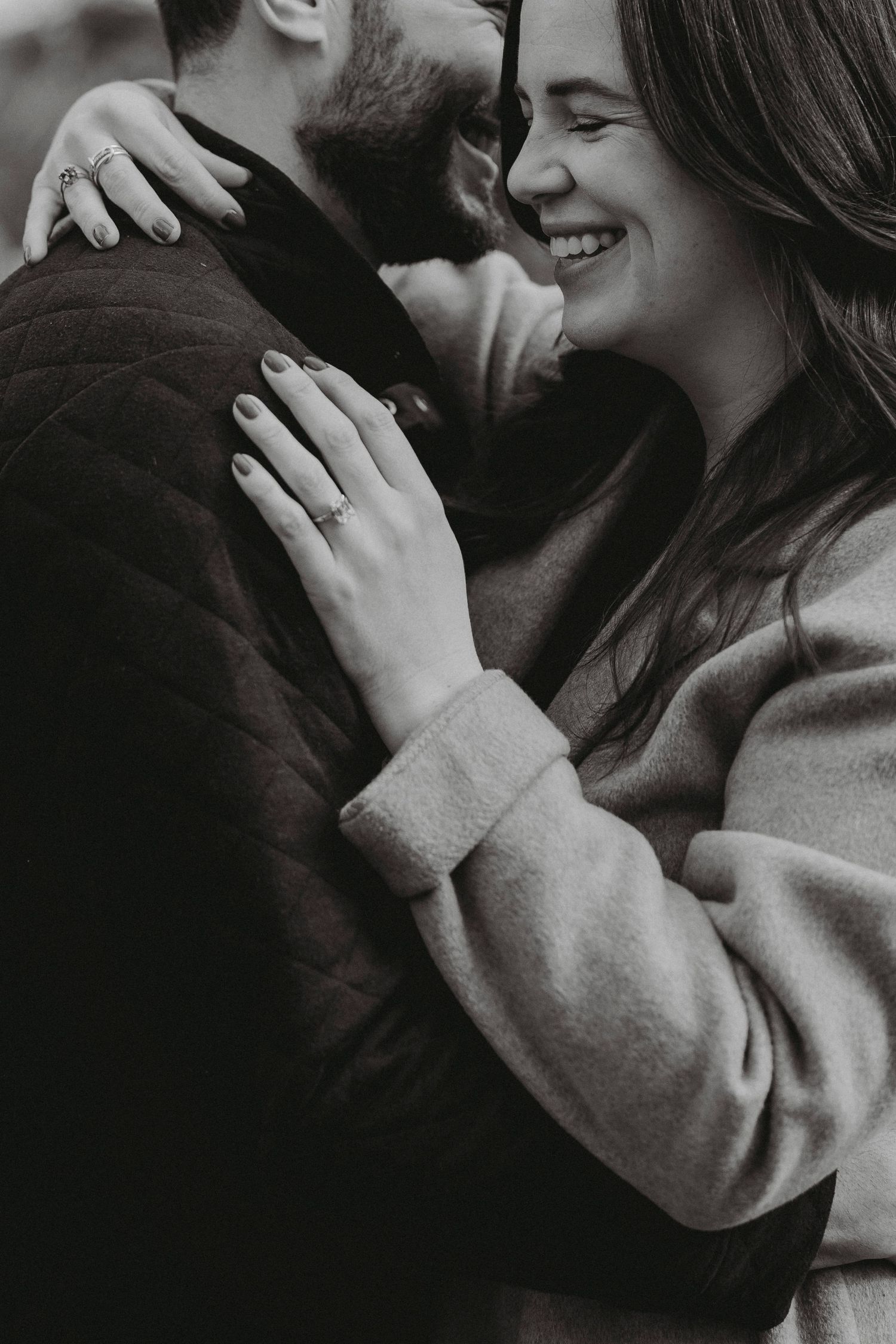 Black and white image of smiling woman embracing man, showing engagement ring on her finger.