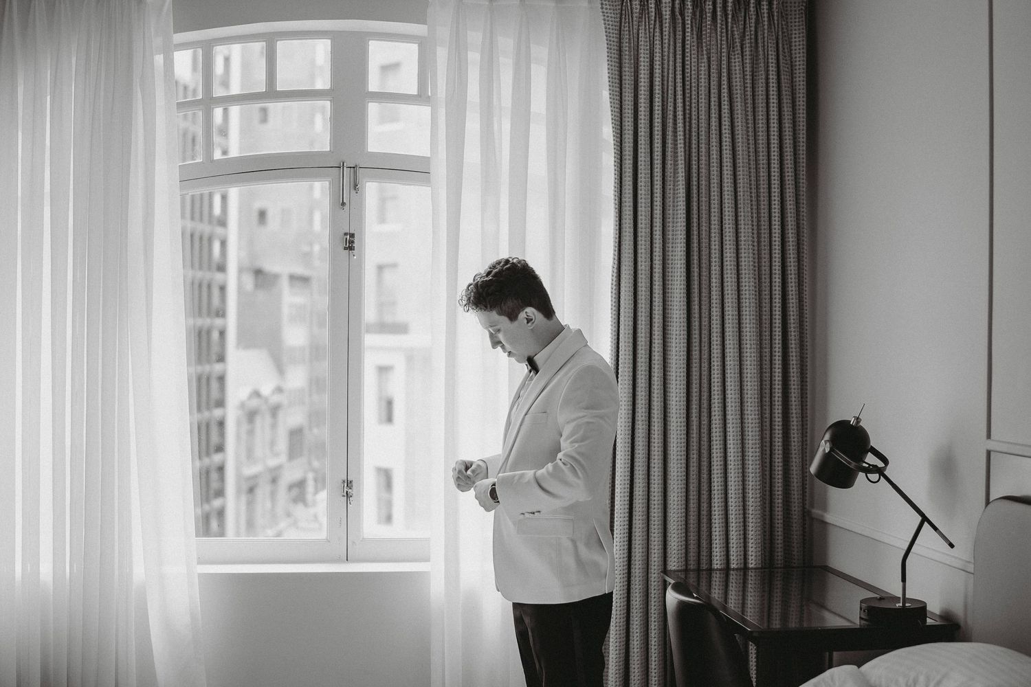 Groom in white jacket adjusting cufflinks by window in black and white wedding preparation photo.