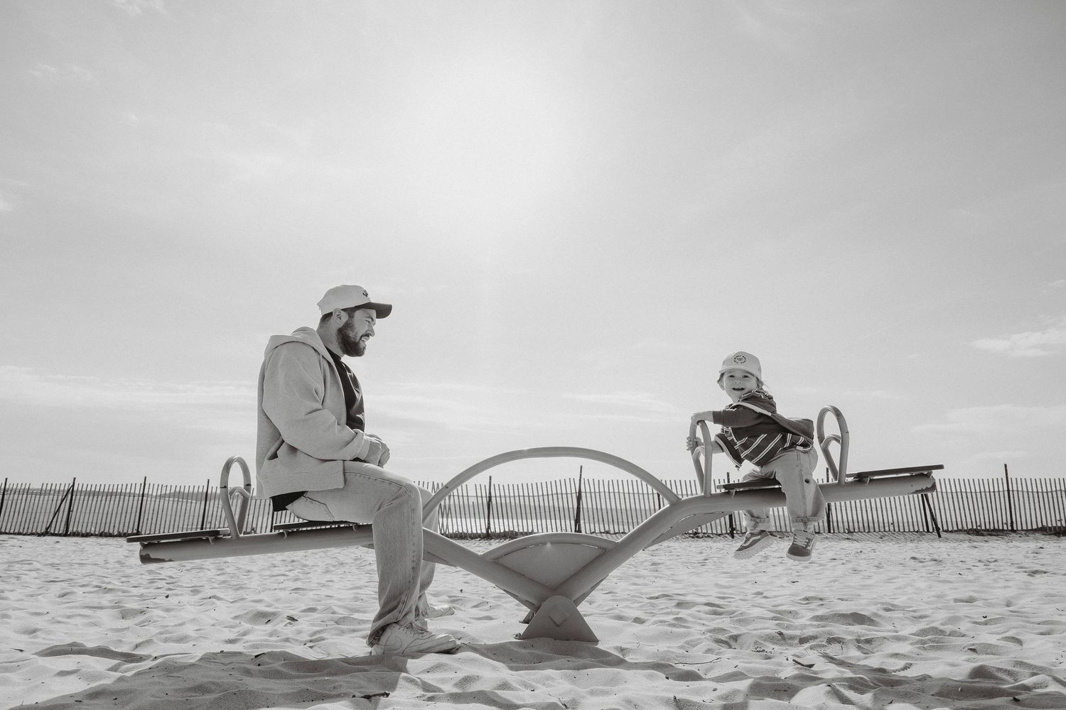 Black and white photo of two people sitting on a seesaw at a sandy beach playground.