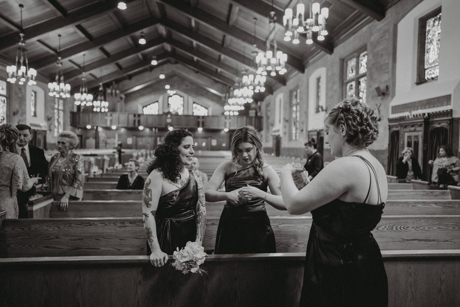 Bridesmaids in black dresses chat inside ornate church with chandeliers before wedding ceremony.