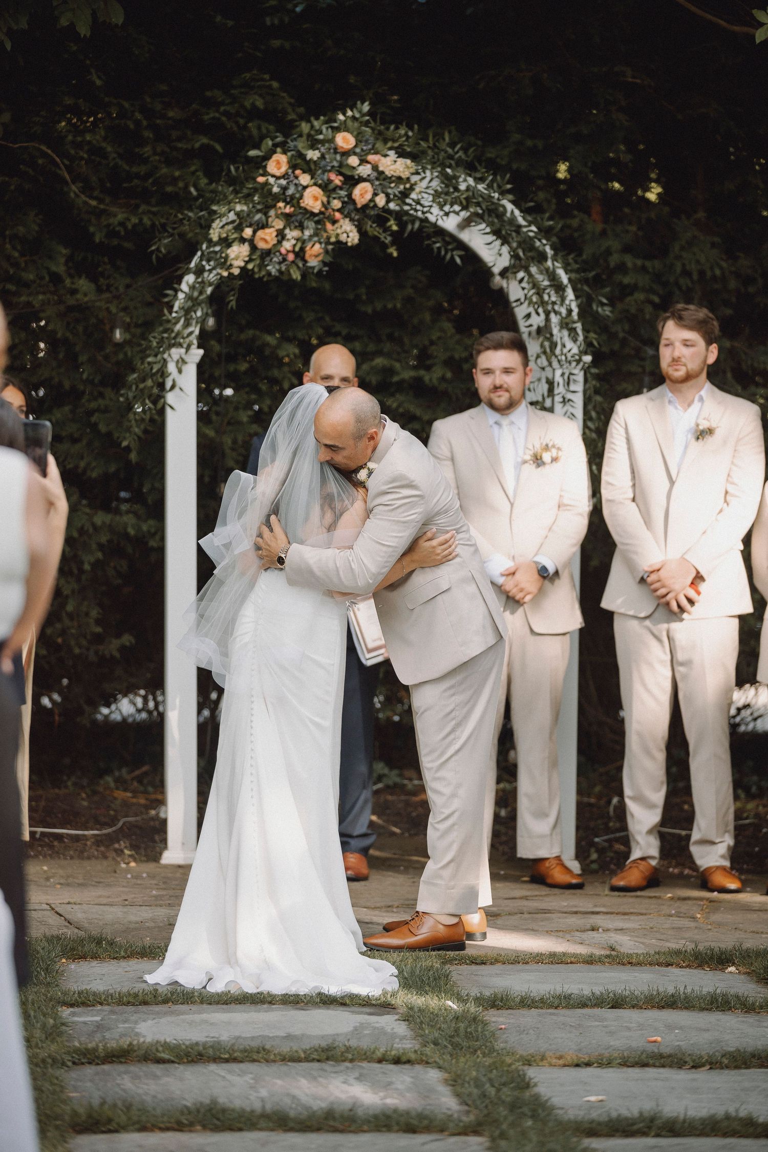 Bride and groom share first kiss under floral arch at outdoor wedding ceremony.