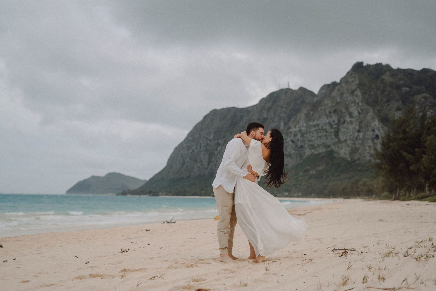 Couple sharing romantic kiss on sandy beach with turquoise ocean and dramatic mountain cliffs behind them.