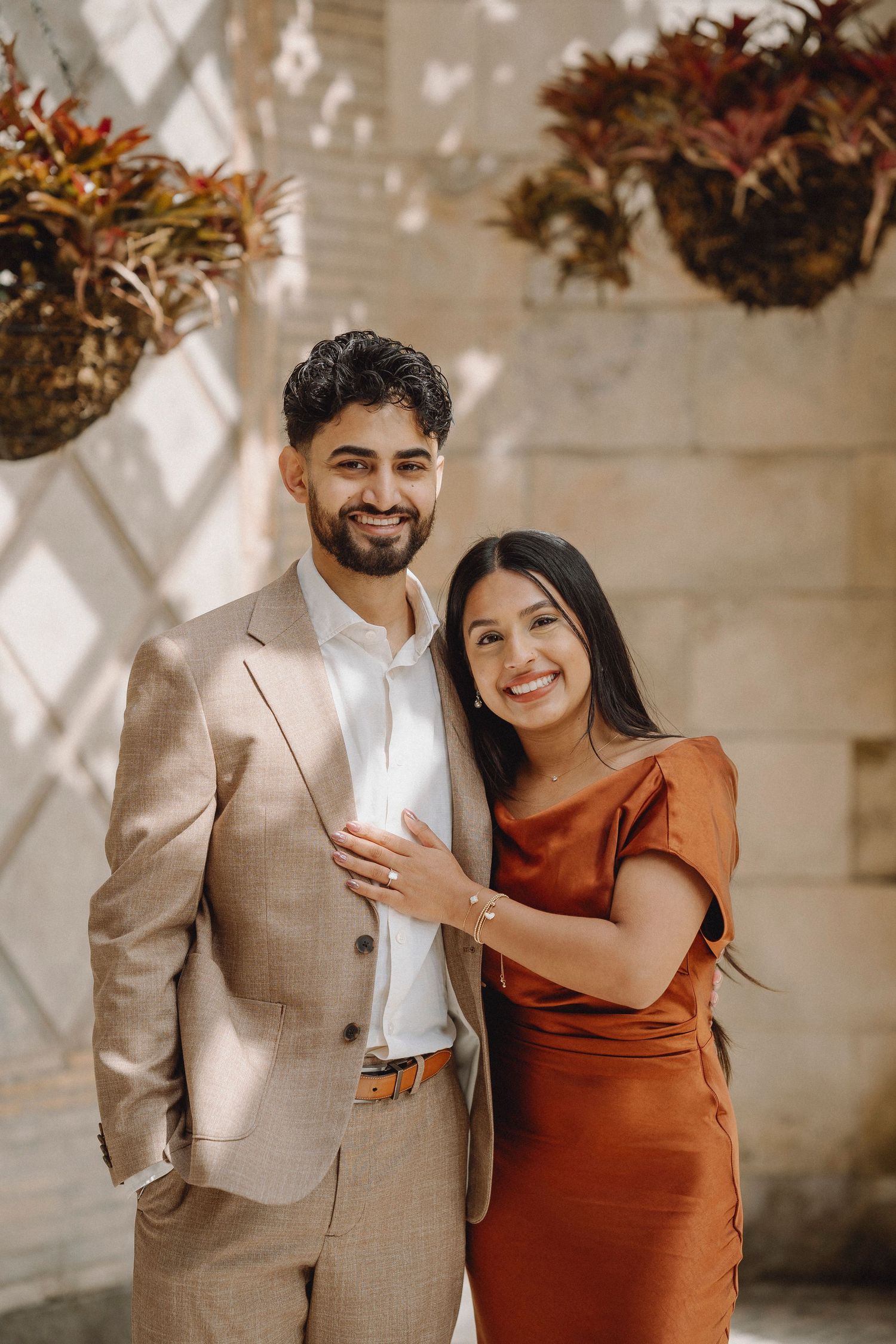 Smiling couple in beige suit and rust dress pose together in a sunlit garden colonnade.