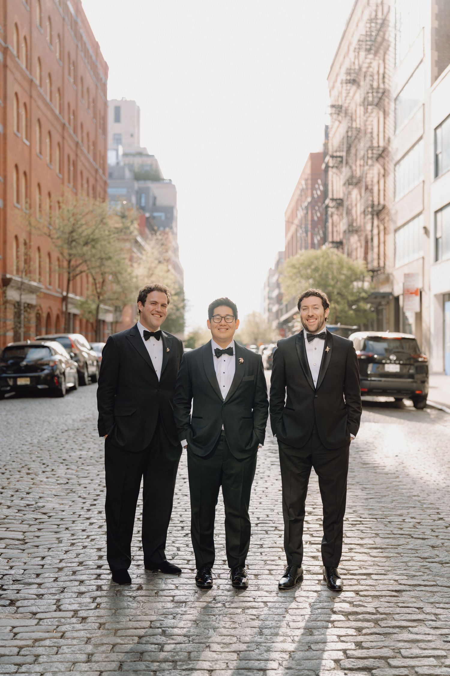 Groom and two groomsmen in black tuxedos pose on cobblestone street in New York City.