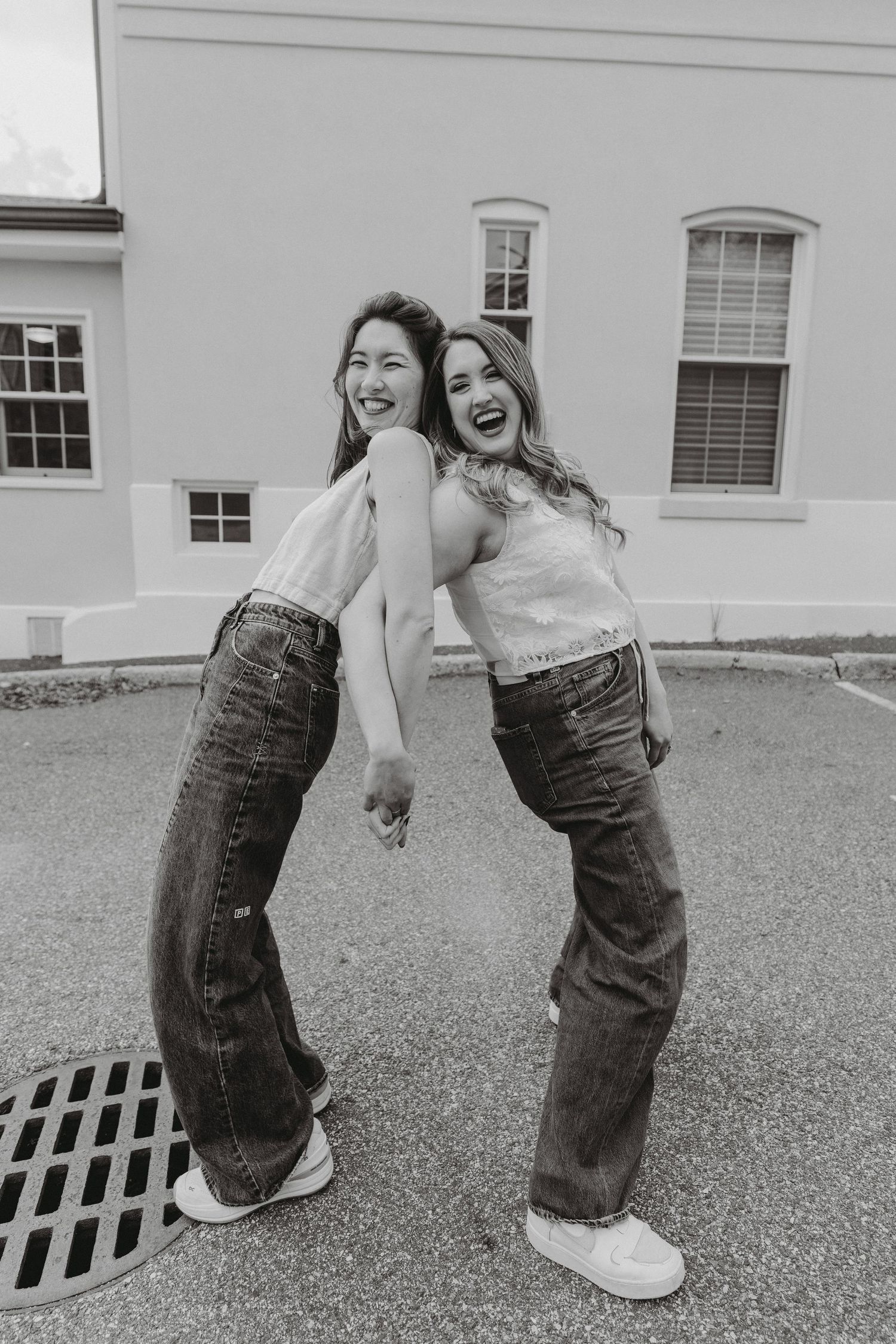 Two laughing women in jeans and white tops pose playfully outside a building in black and white photo.