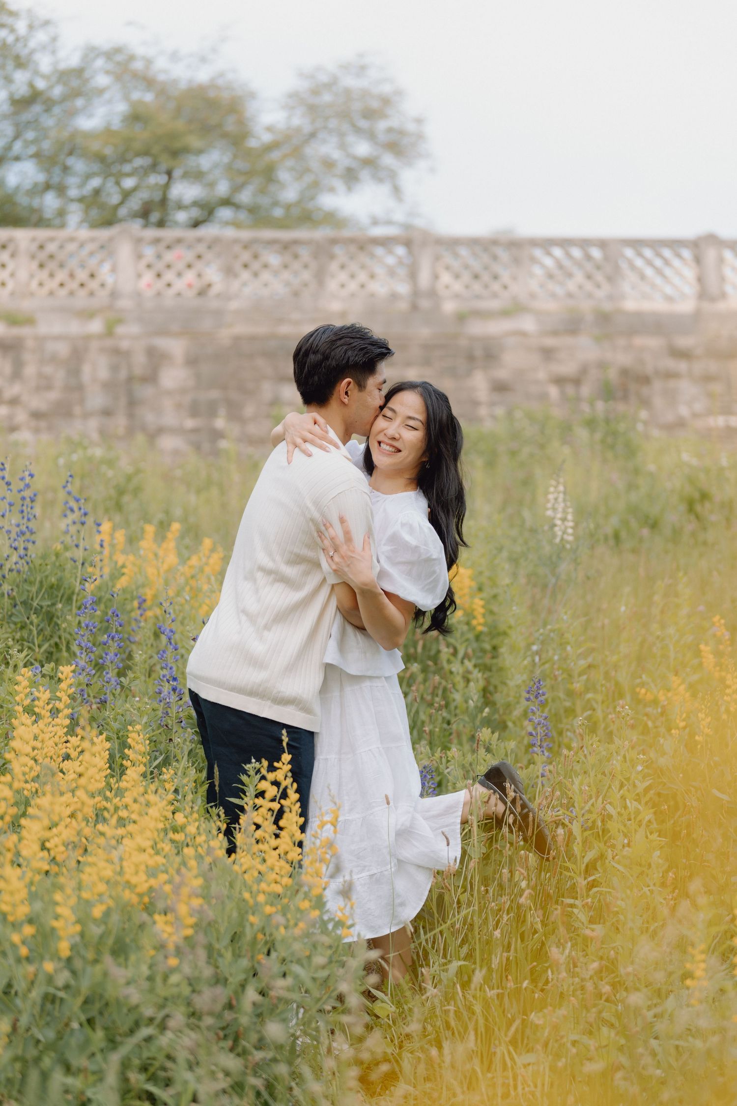 Smiling couple embracing in a sunlit meadow of yellow and purple lupine wildflowers with a stone balustrade in the background.
