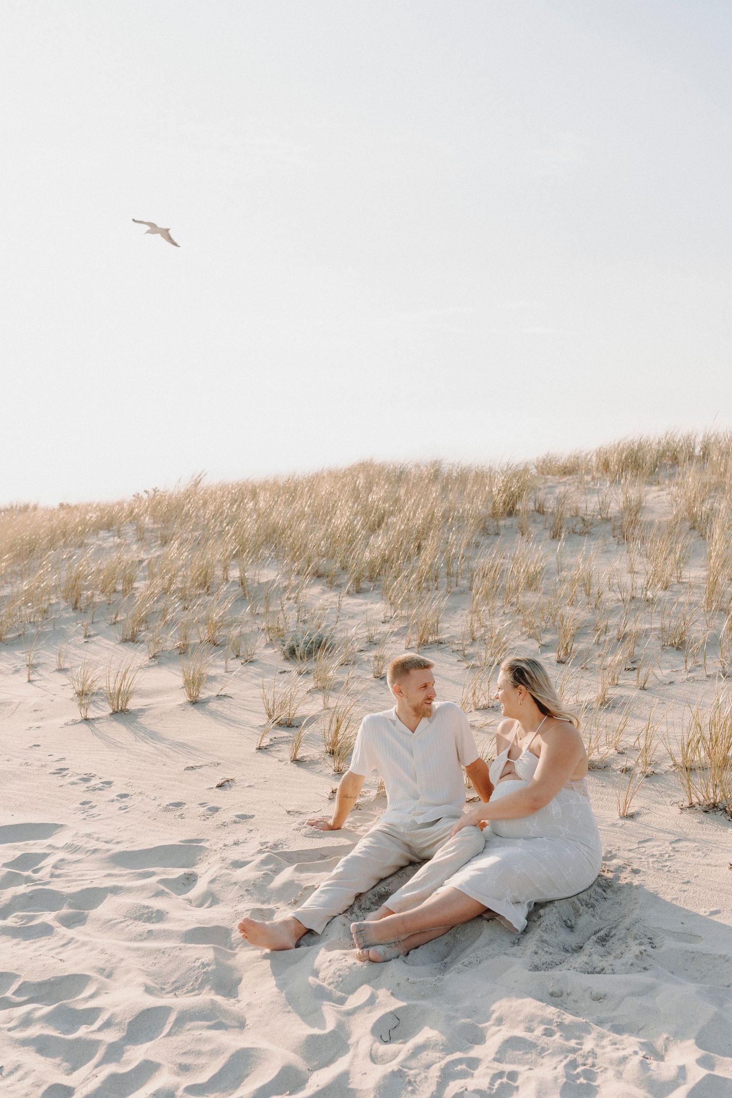 Happy couple sitting on sandy beach dunes sharing drinks in golden sunlight.